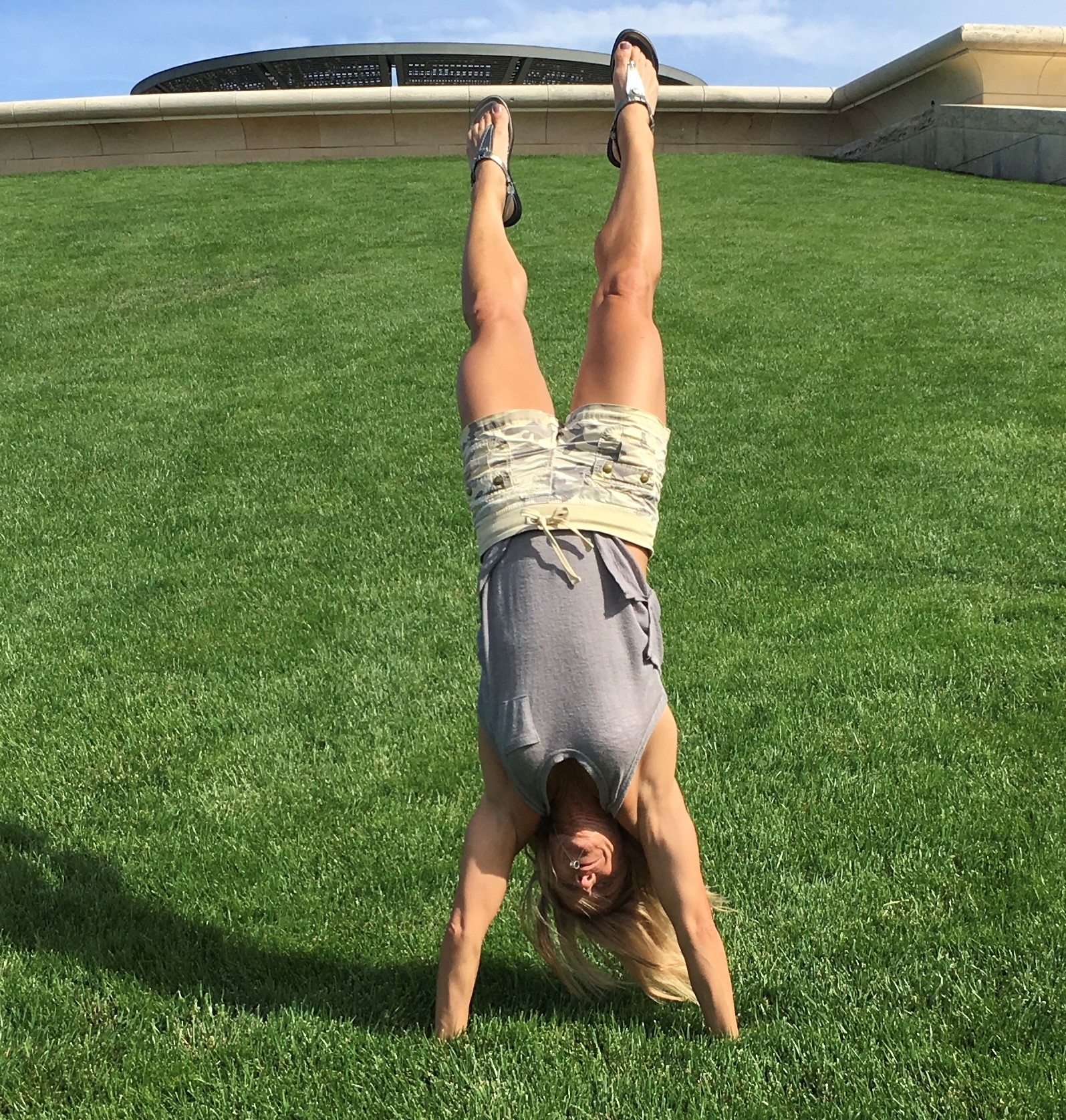 A woman does a handstand on the grass at the bottom of a hill. There’s a building behind a wall at the top of the hill.