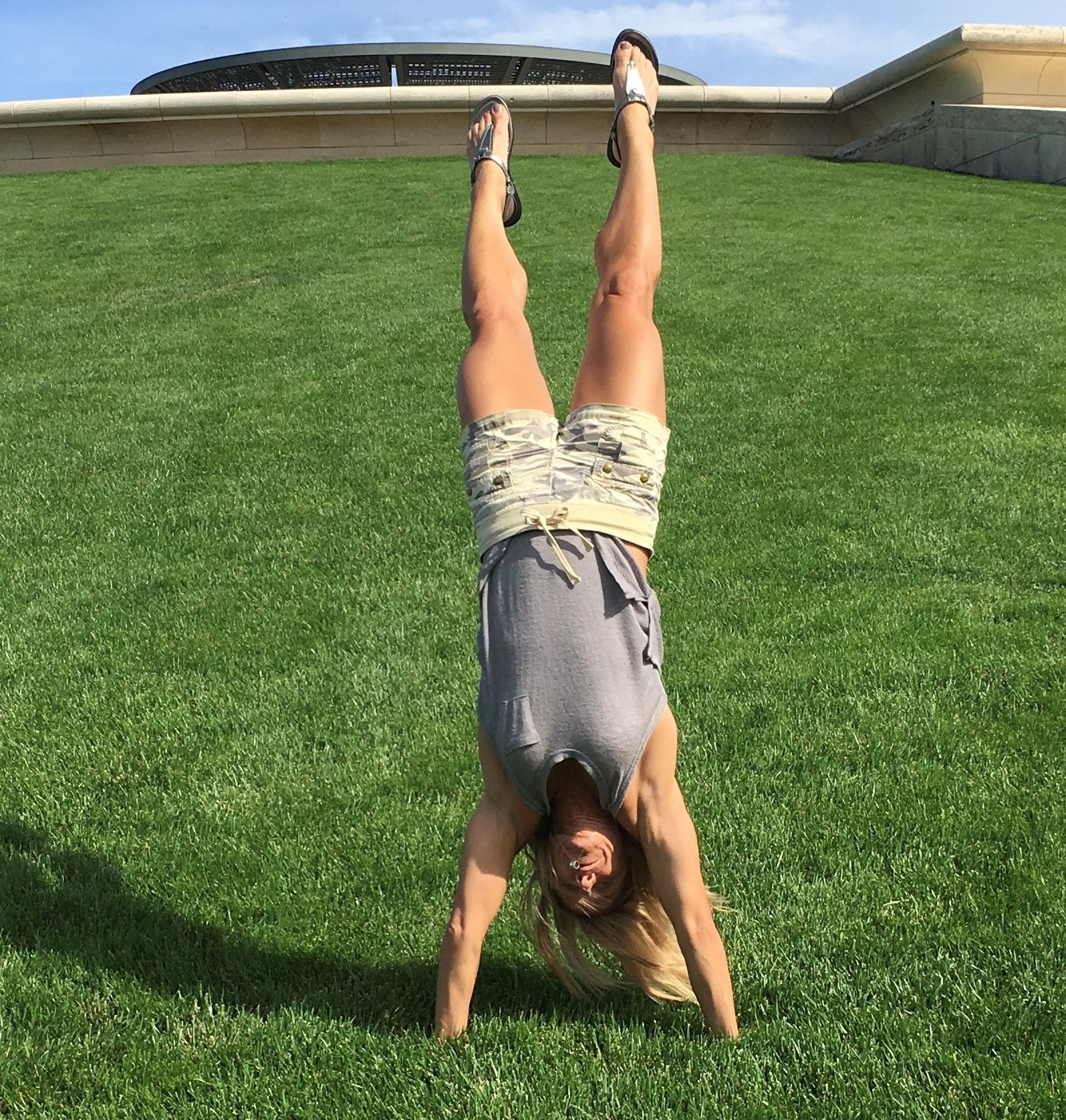 A woman does a handstand on the grass at the bottom of a hill. There’s a building behind a wall at the top of the hill.