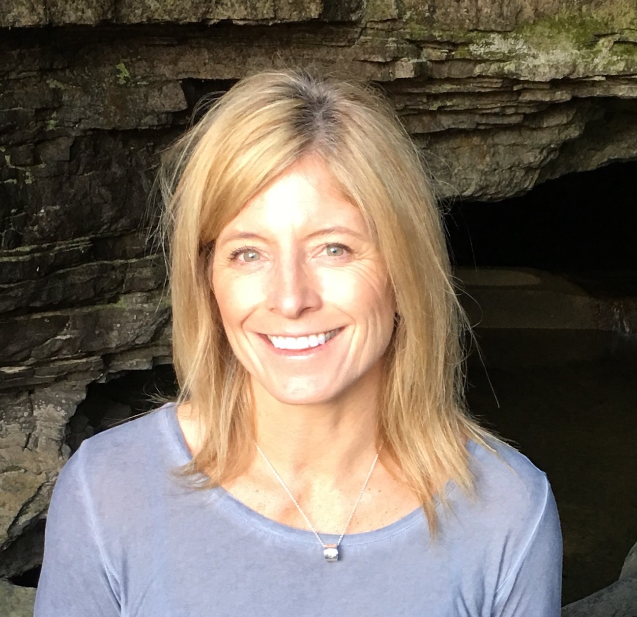 A profile photo of a smiling middle aged woman who has golden brown hair and is wearing a blue casual shirt and a necklace.