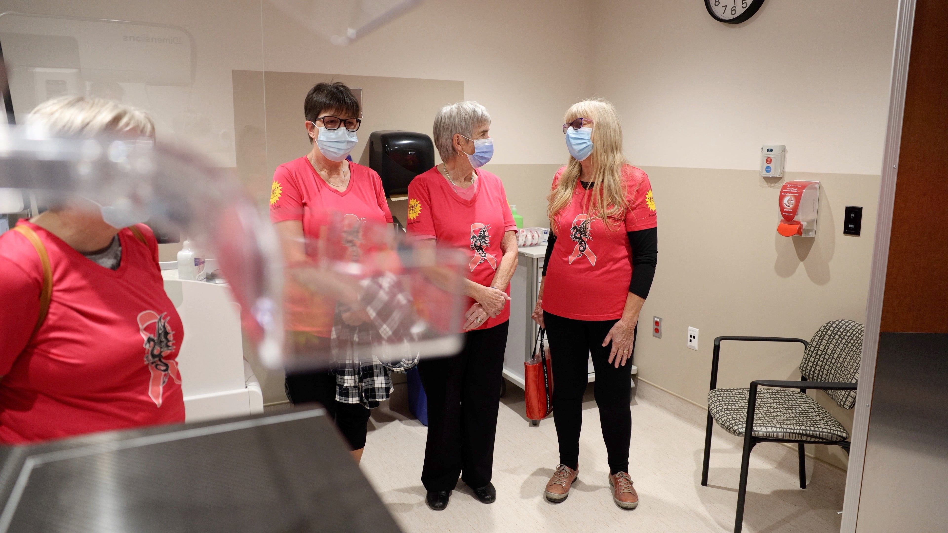 Three people wearing pink shirts and masks stand and talk inside.