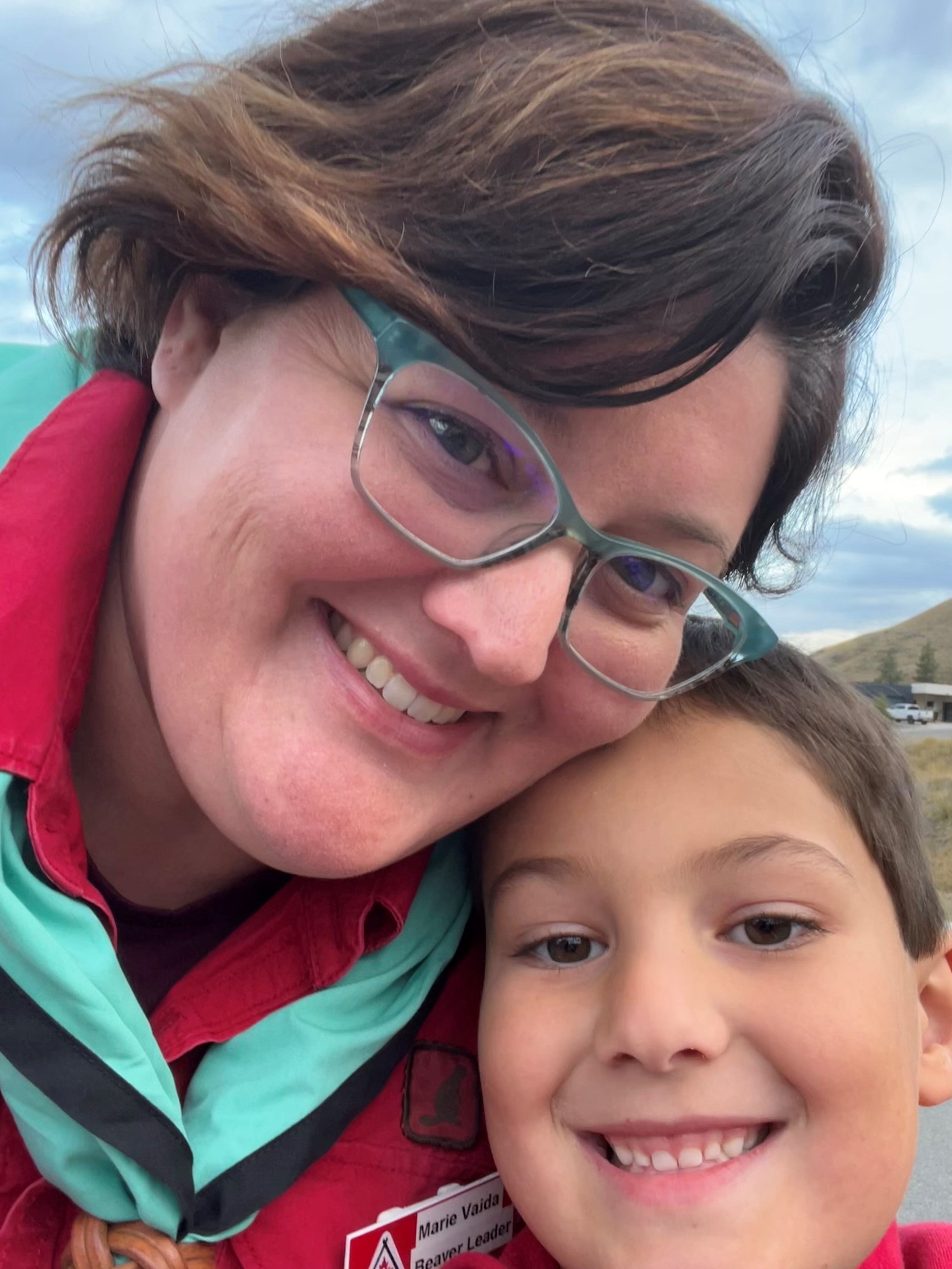 A smiling woman in glasses wearing a red shirt and teal scarf cuddles a young boy