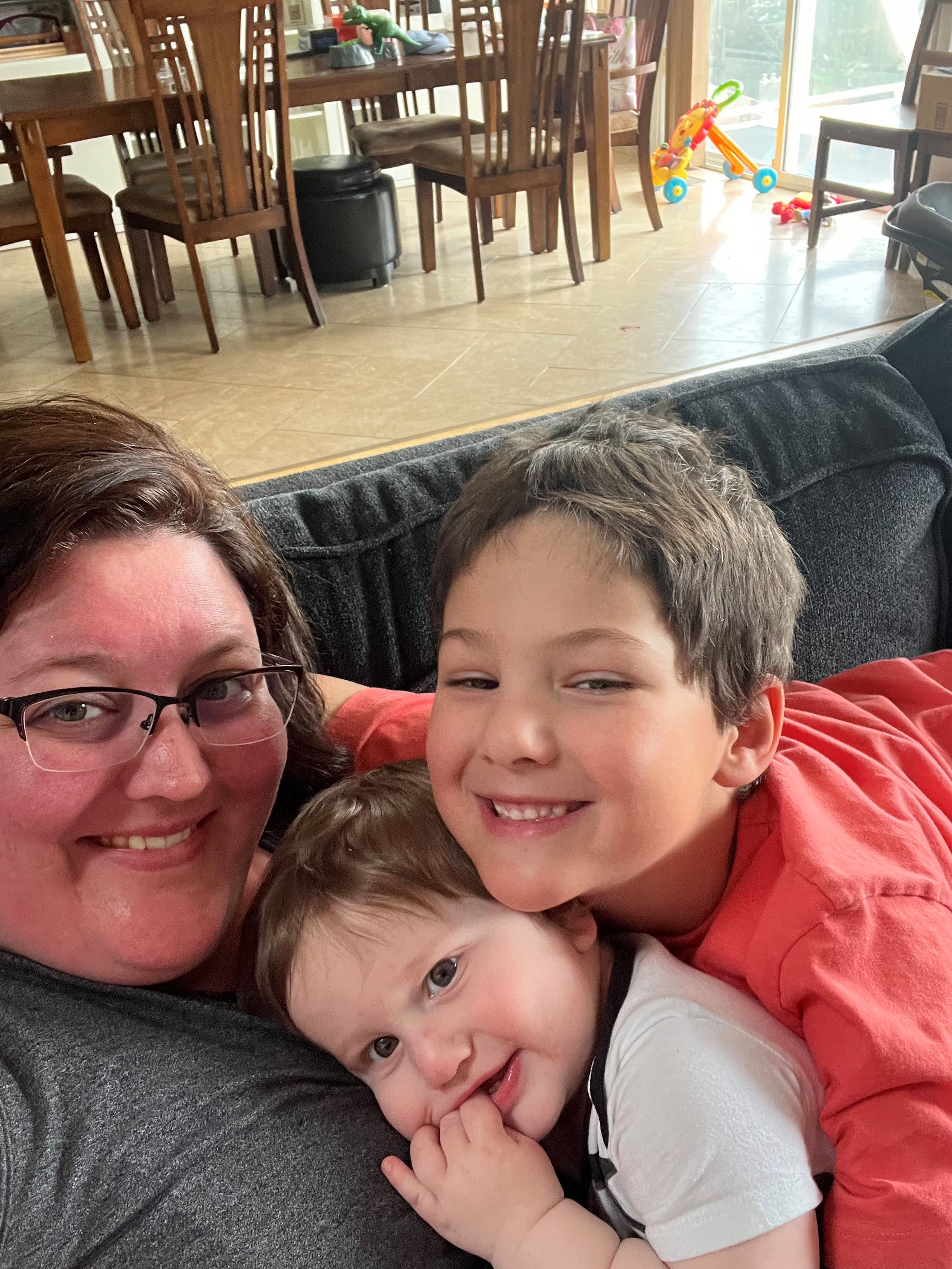 A smiling woman with glasses and grey shirt sits on a grey couch cuddling two young boys