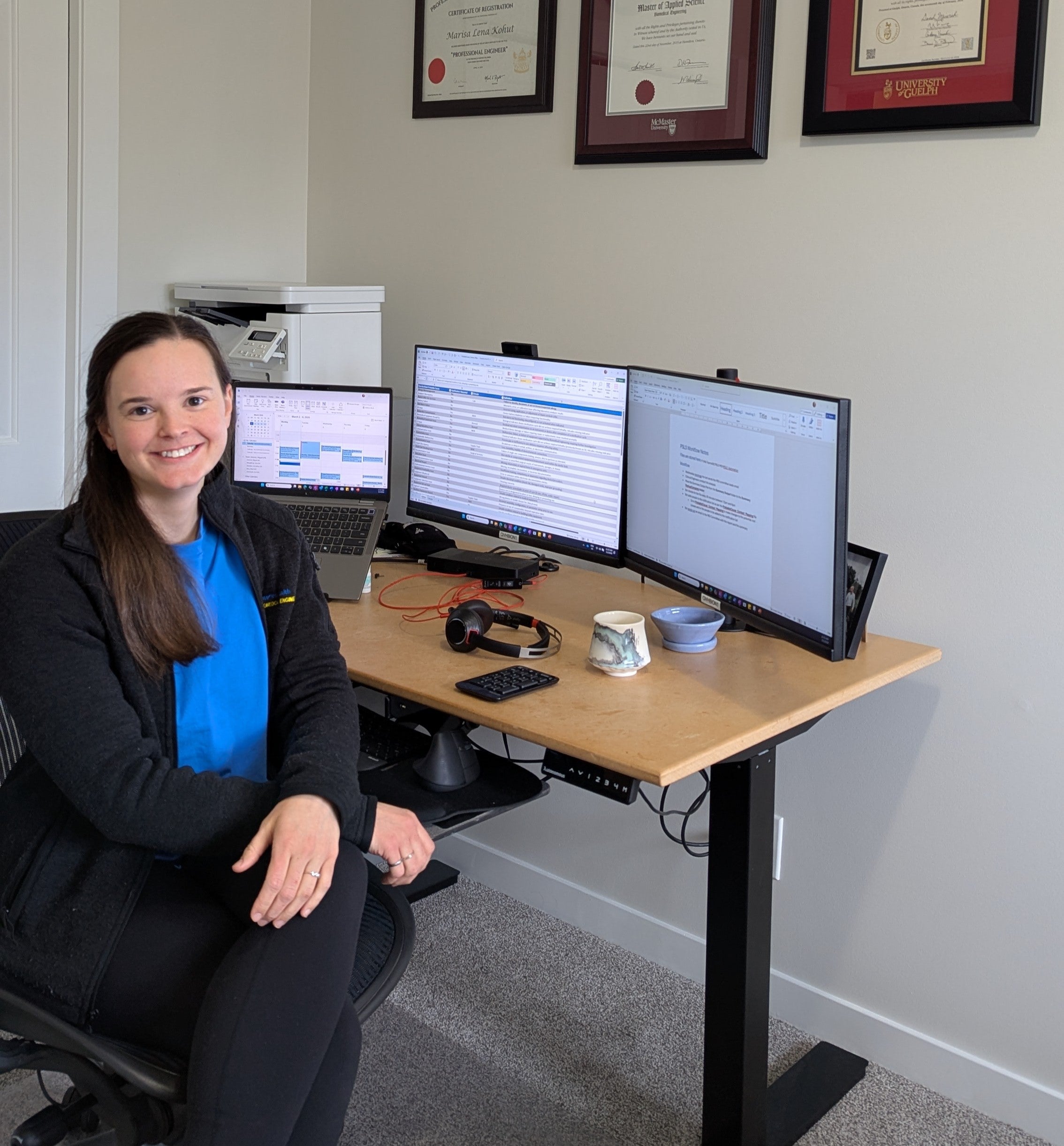 A person in a blue shirt and black jacket sits at a workstation with three monitors.