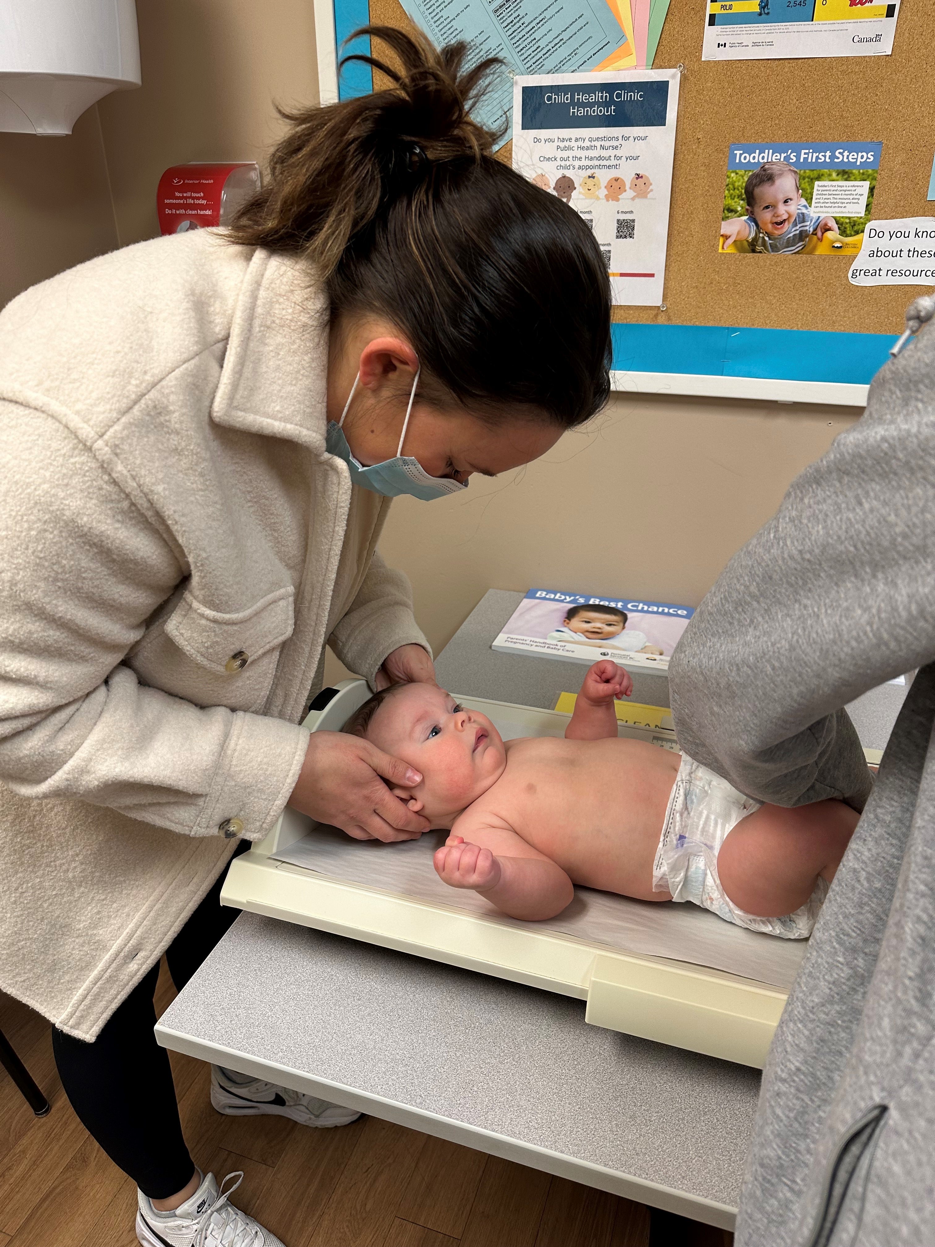 Mom with brown hair tied back, wearing white jacket, soothing baby who is laying down for medical examination.