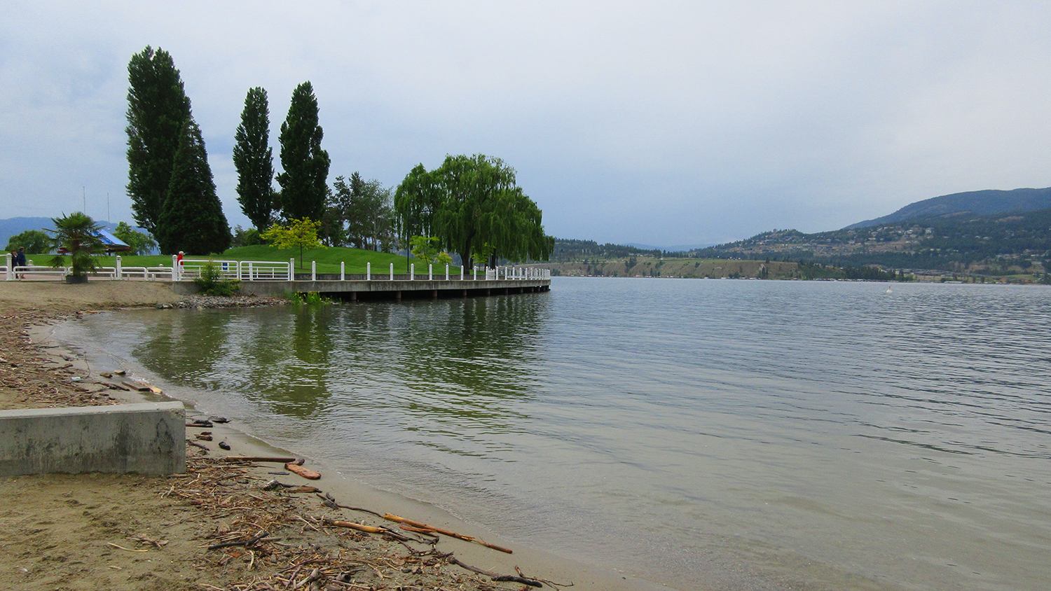A nature shot of a body of water with a beach, park and walkway to the left and forested mountains in the background.