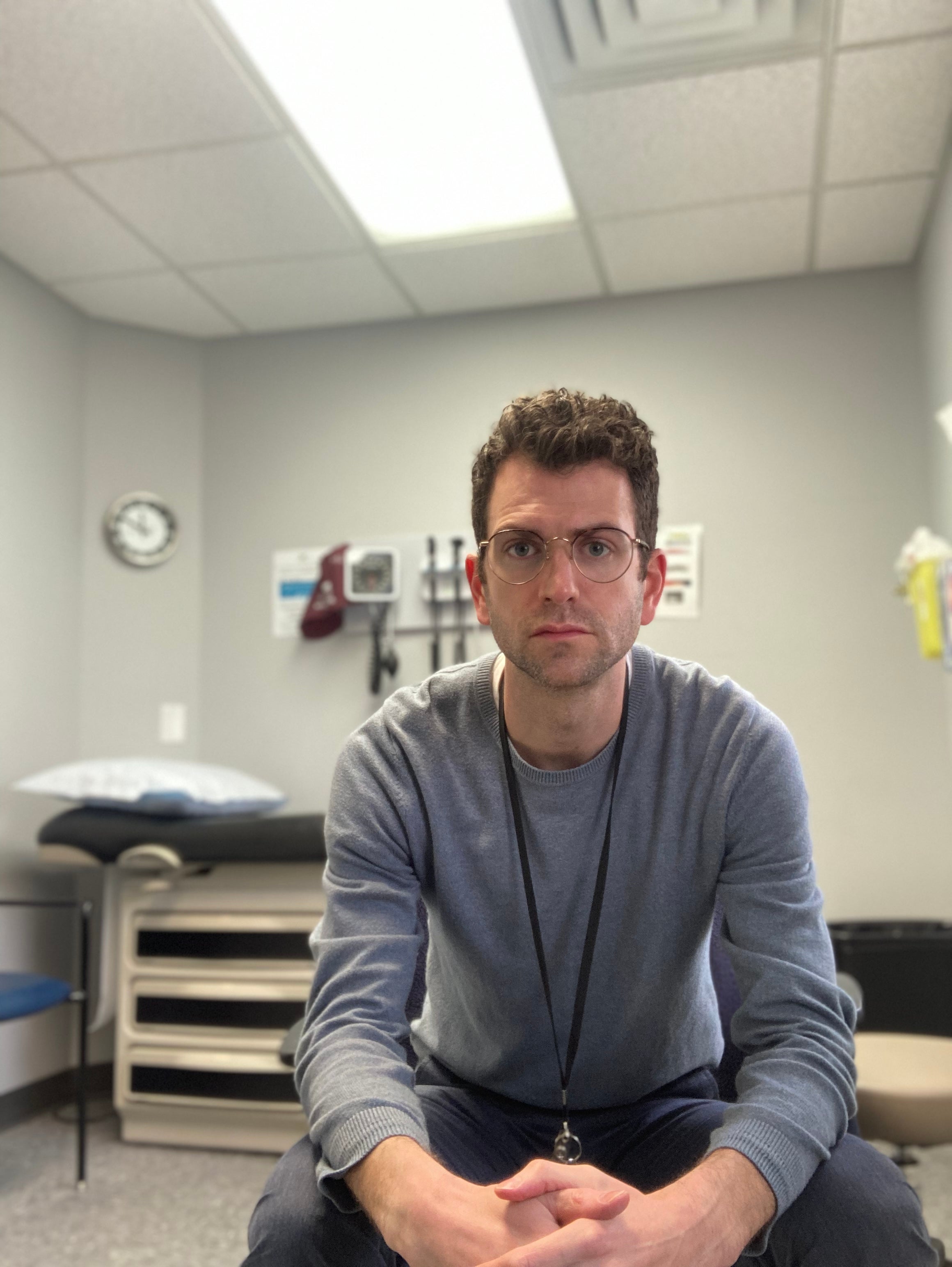 A person in a grey long-sleeved shirt with hands clasped sits forward on a chair in a medical room with a fluorescent light overhead,