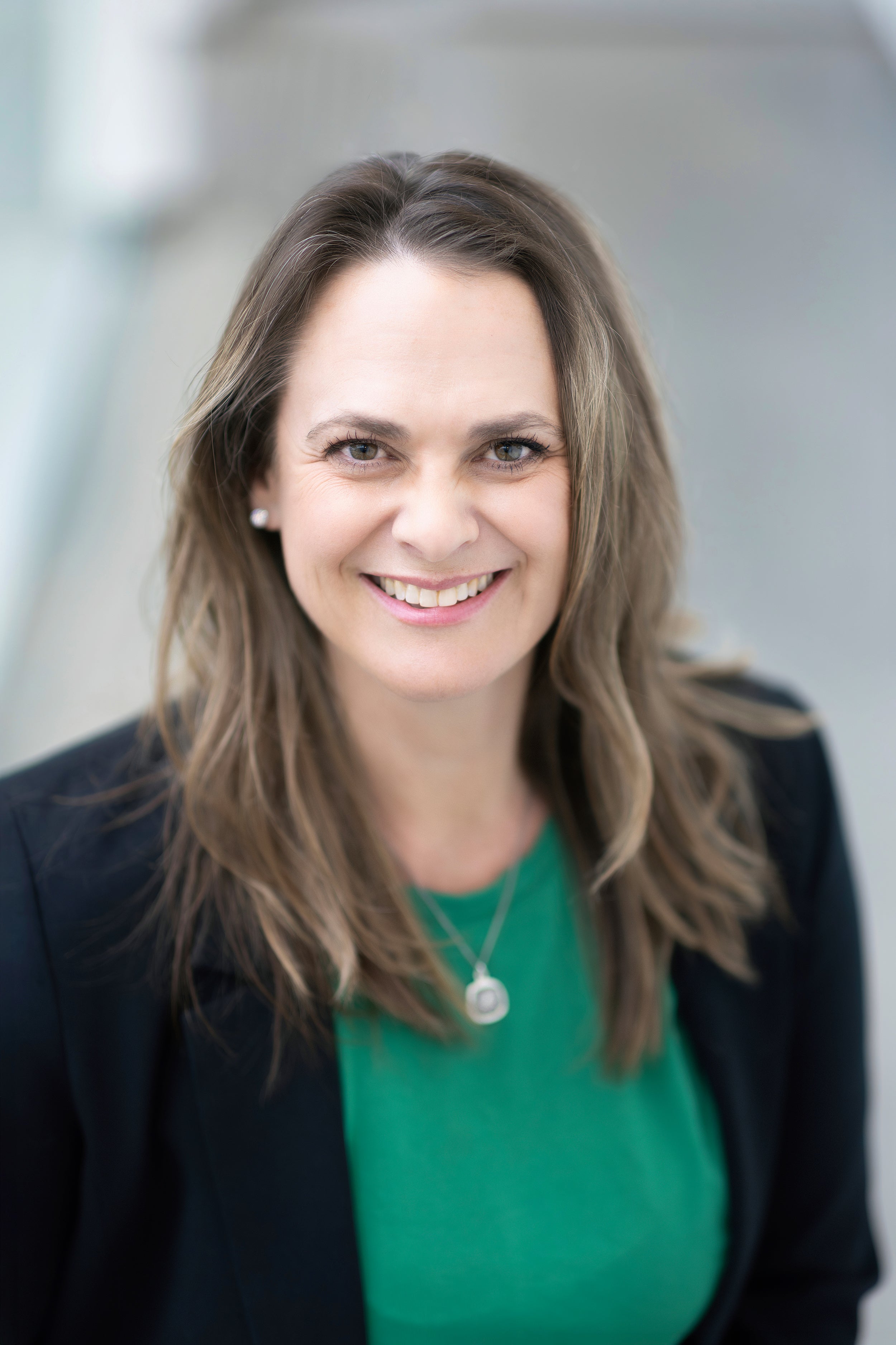 Profile shot of a smiling person with long brown hair wearing a necklace over a green shirt with a black blazer.