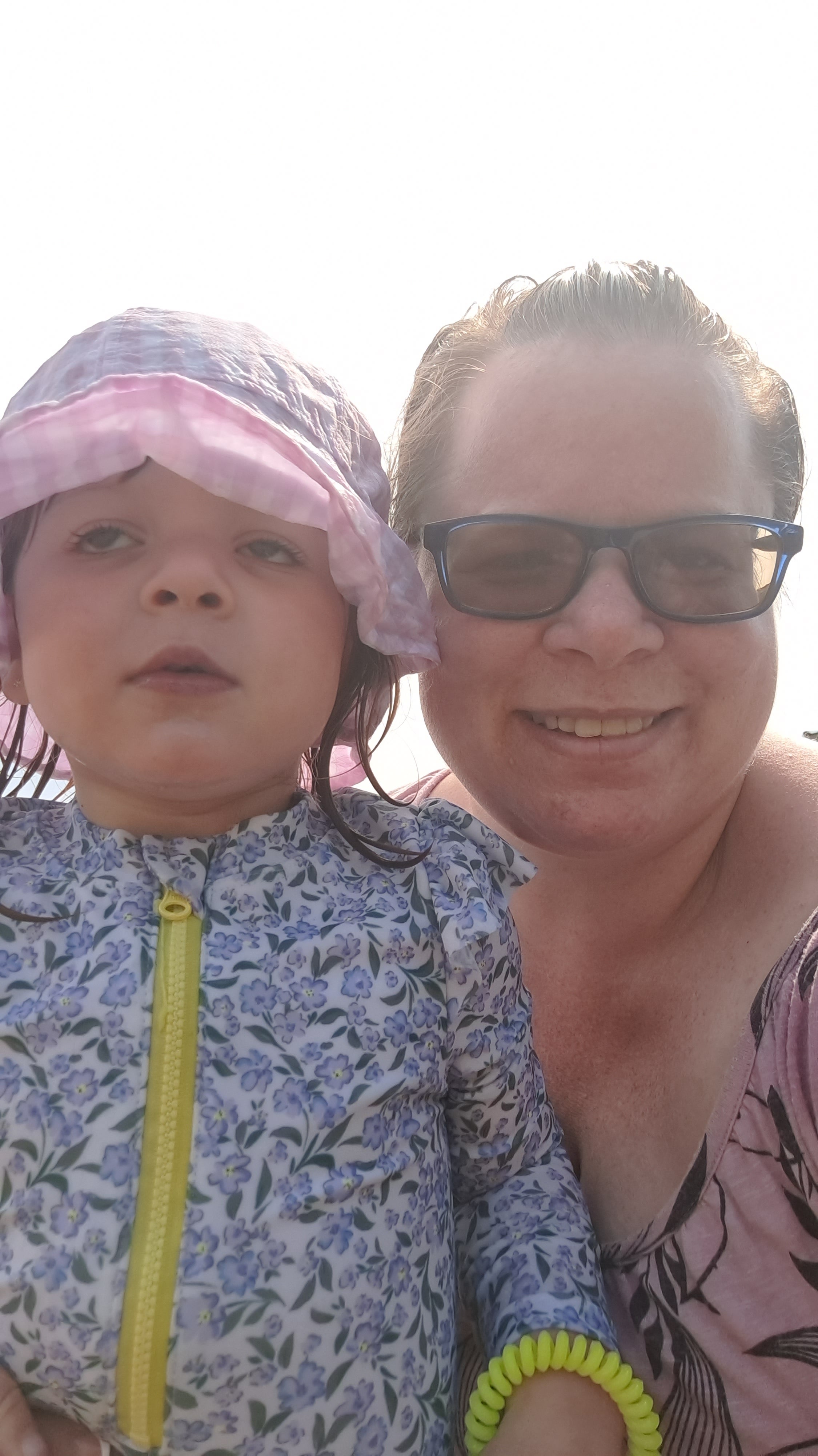 A selfie of a woman wearing sunglasses who has a young girl on her lap who is wearing a coat and hat.