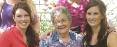 Two people both with long brown hair stand on either side of an elderly person wearing glasses and a purple blouse.