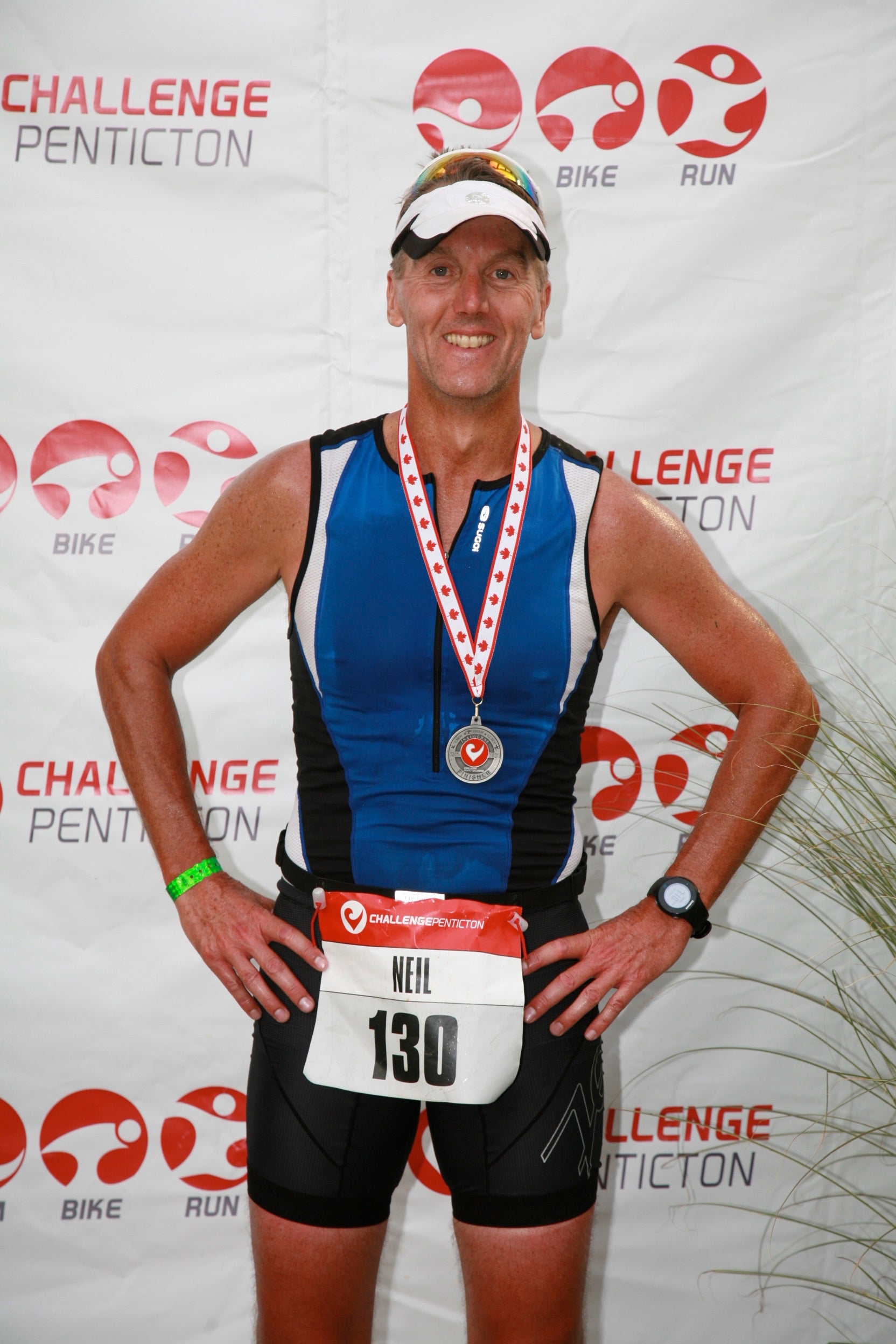 A participant wearing a triathlon racing kit and medal poses in front of a Challenge Penticton event backdrop.