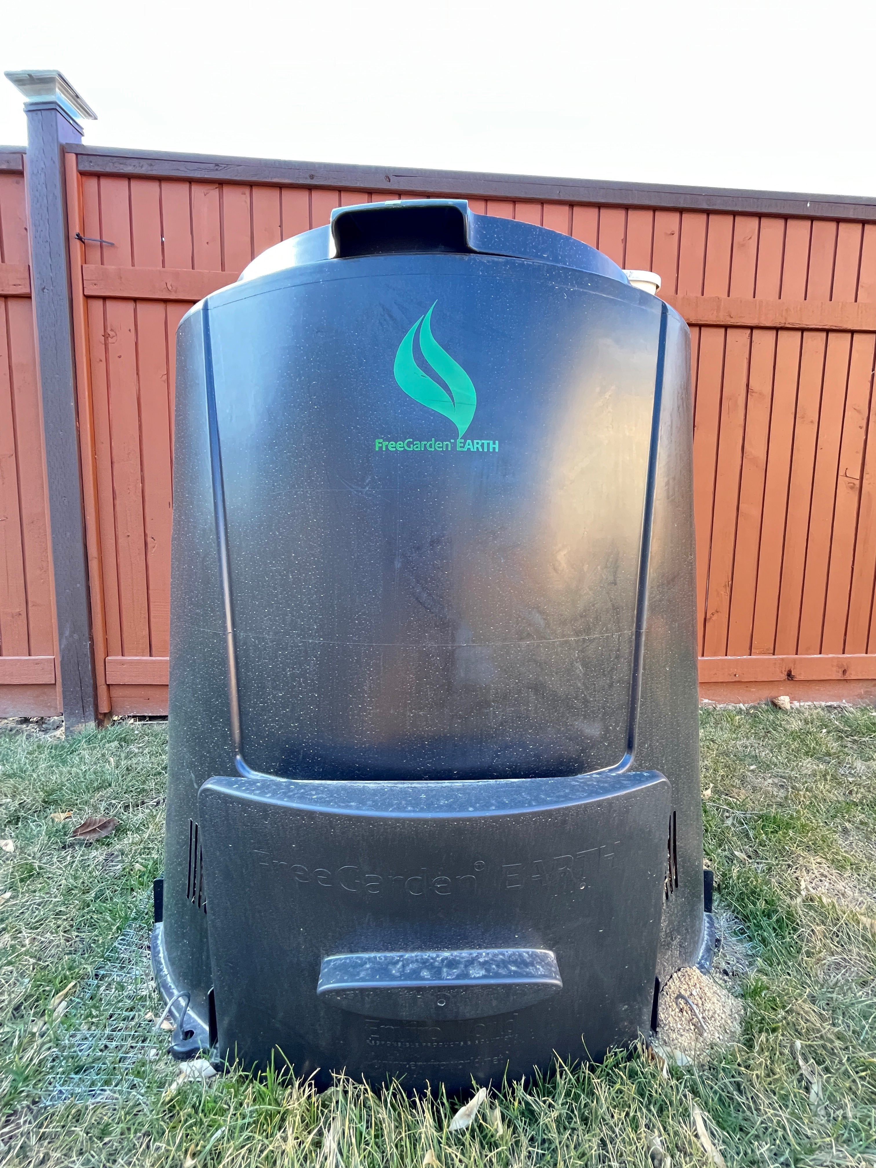 A black compost bin with a green leaf image sits on green grass in front of an orange fence