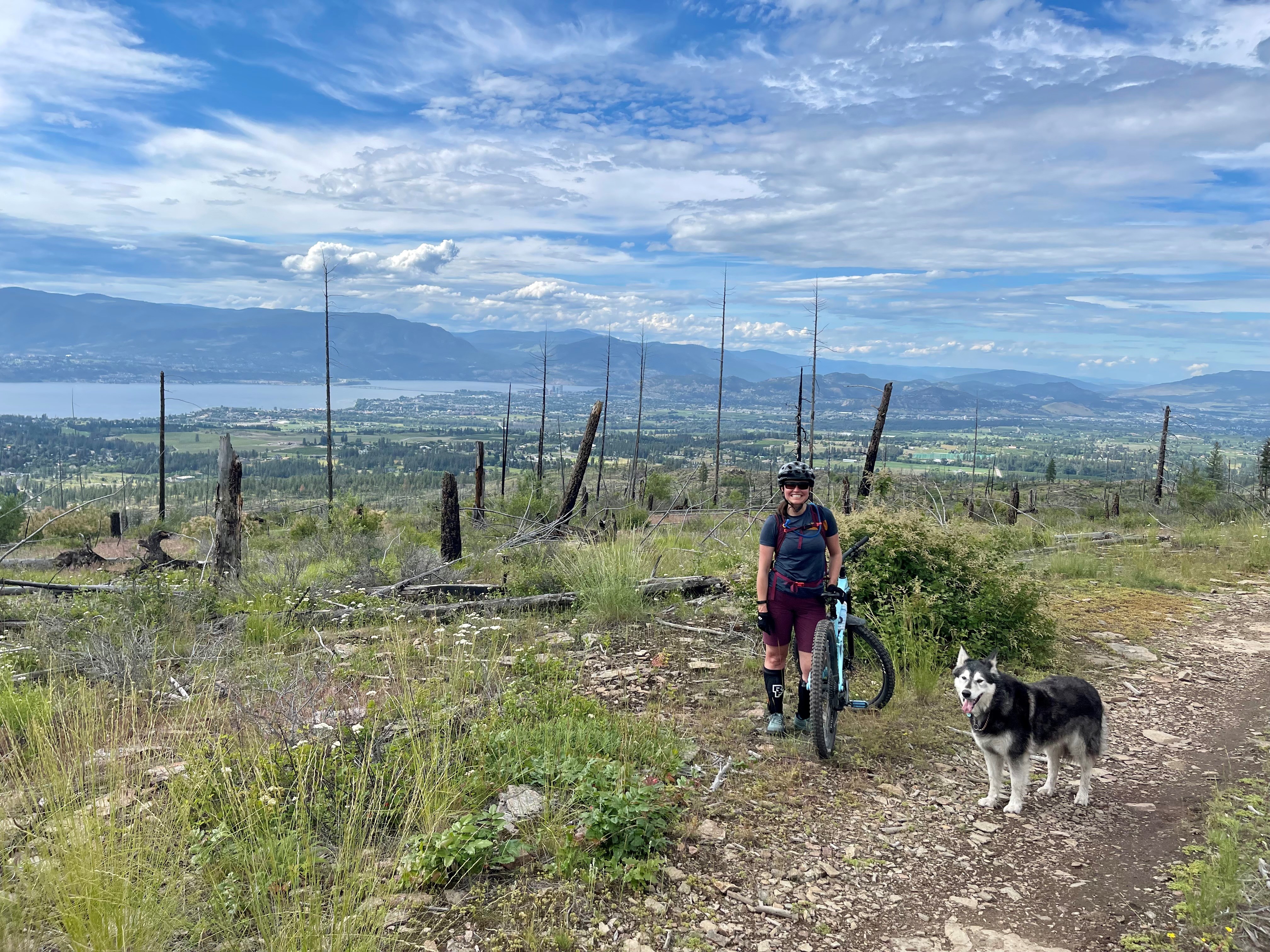 A woman poses on her mountain bike alongside her dog on a trail.