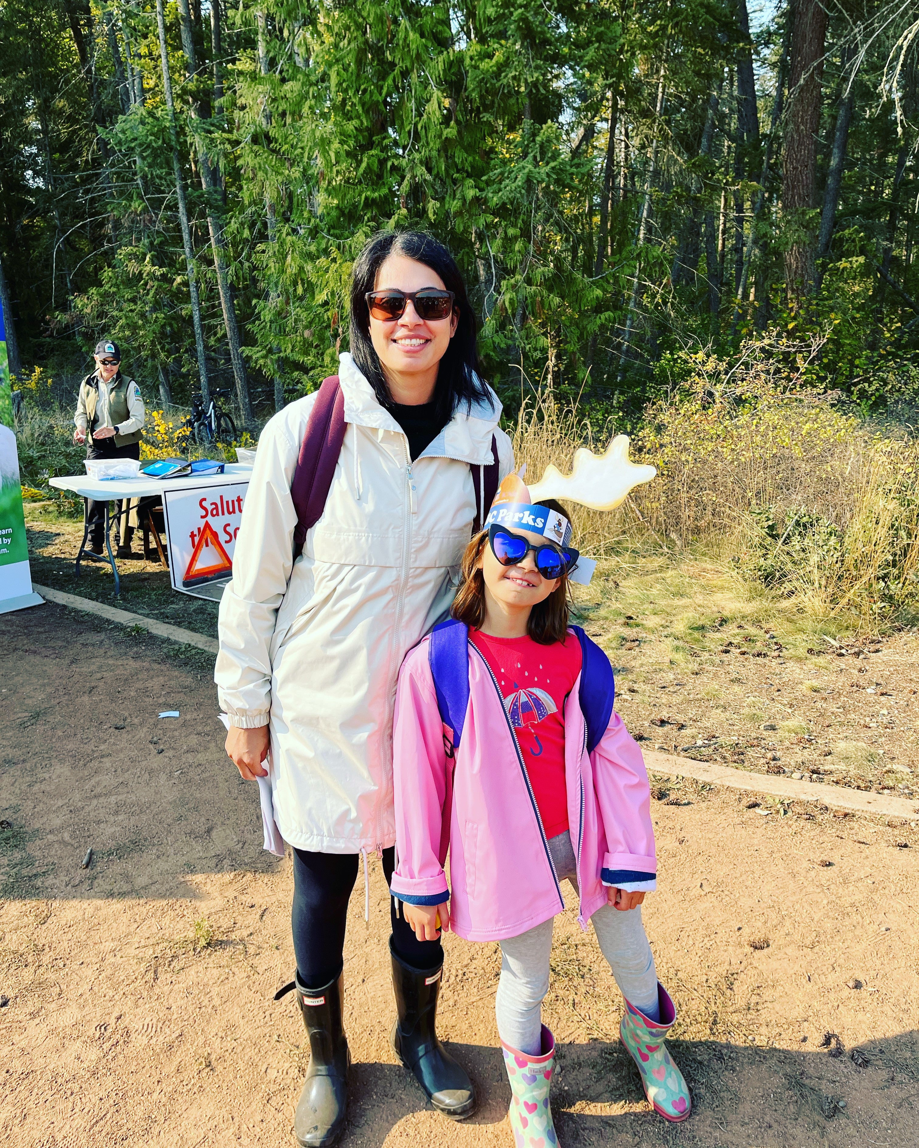 A woman and child in fall gear and wearing sunglasses stand by the forest with a promotional booth visible in the background