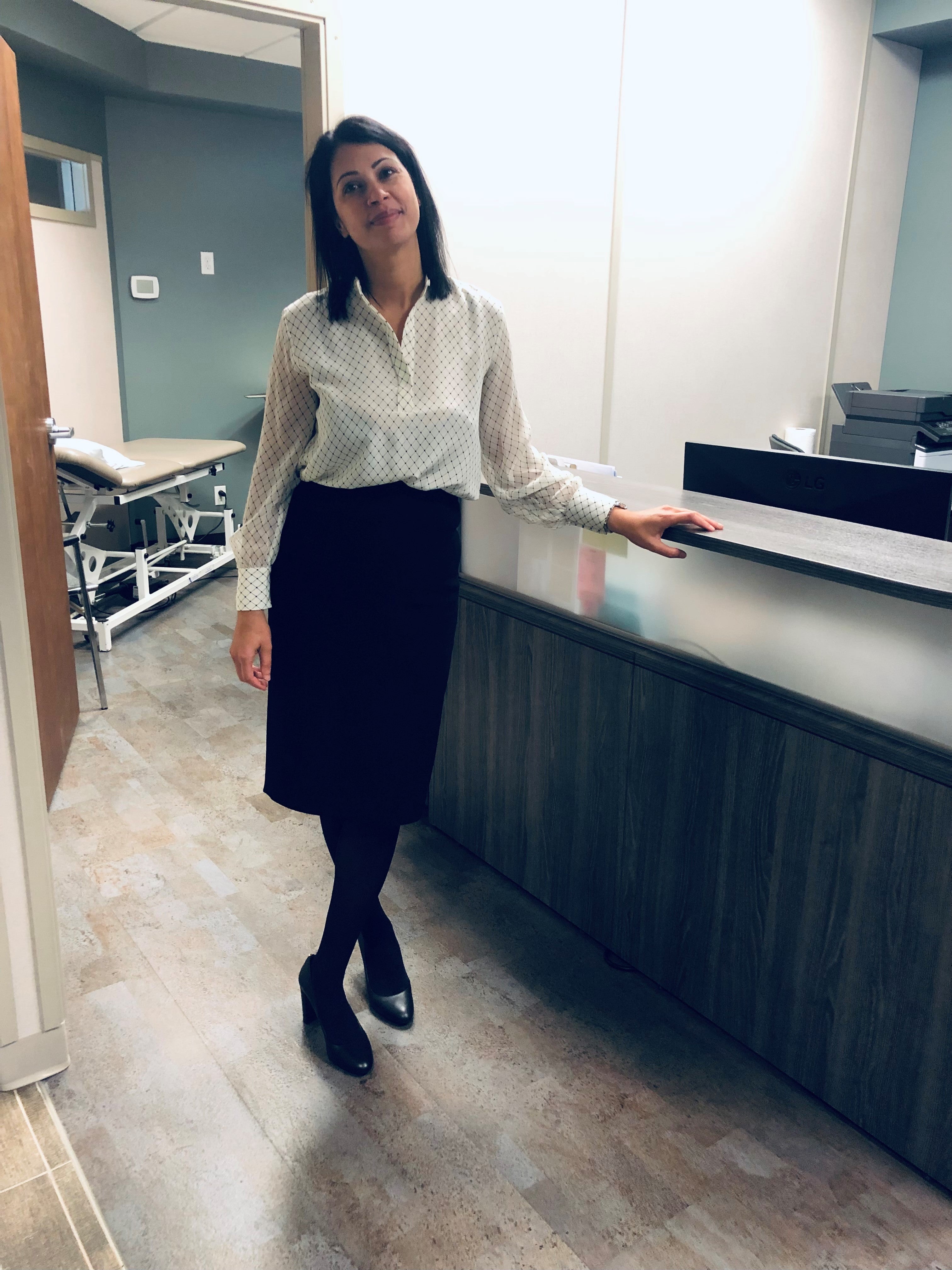 A woman in business attire stand with her hand on a reception desk. An examination bed can be seen through an open door in the background