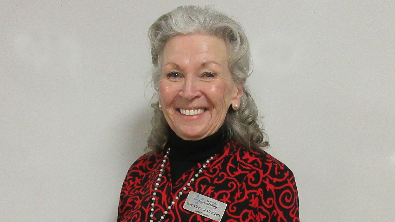 A smiling person with silver-coloured hair poses for a picture. They're wearing a patterned black and red shirt and a pearl necklace.