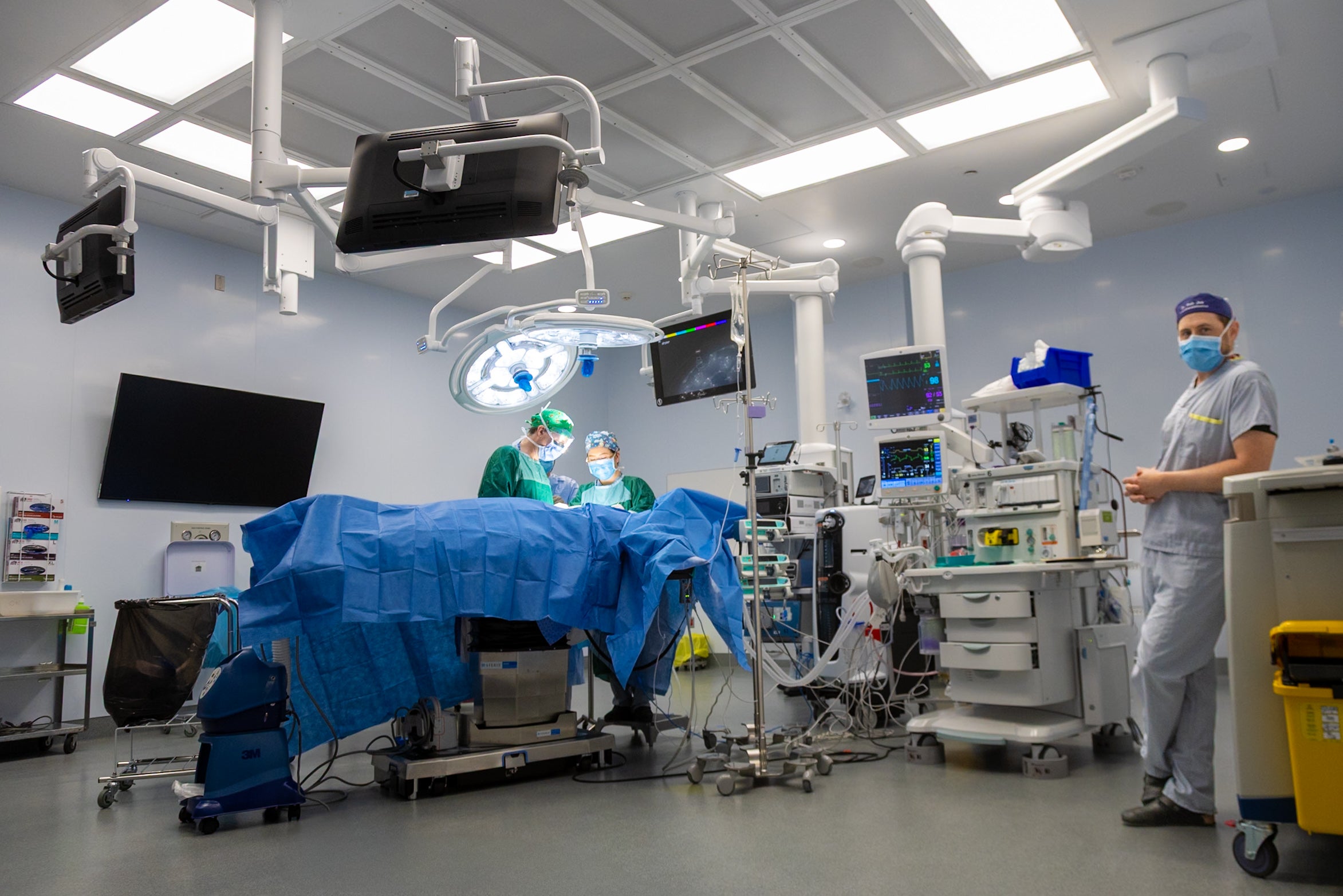 A health-care professional leans against a counter in an operating room as two others perform surgery. The room is full of high tech equipment.