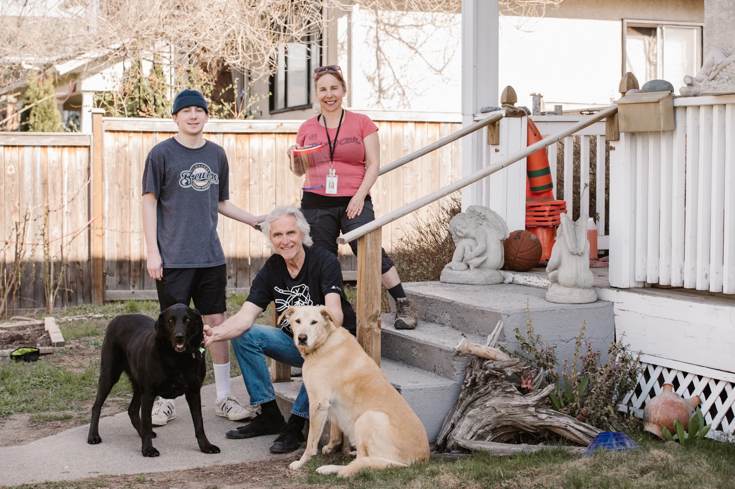 A man with longish white hair wearing a black tshirt, a youth wearing a grey toque and tshirt and a woman wearing a pink tshirt and holding a face shield stand on a porch with two dogs, one black and one tan