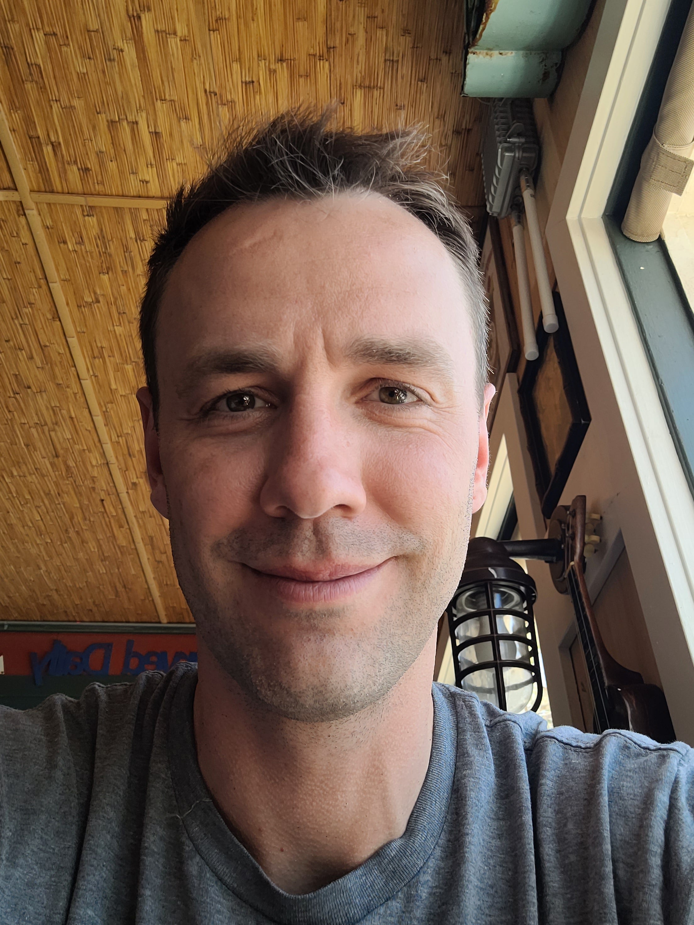 A smiling man with short brown hair wearing a grey tshirt takes a selfie in front of wood panelling