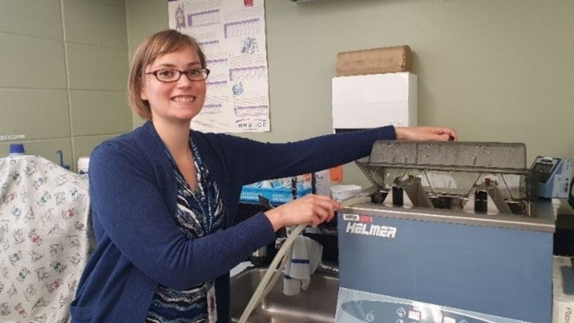 A person with short blond hair and glasses wearing a blue sweater demonstrates how a machine works.