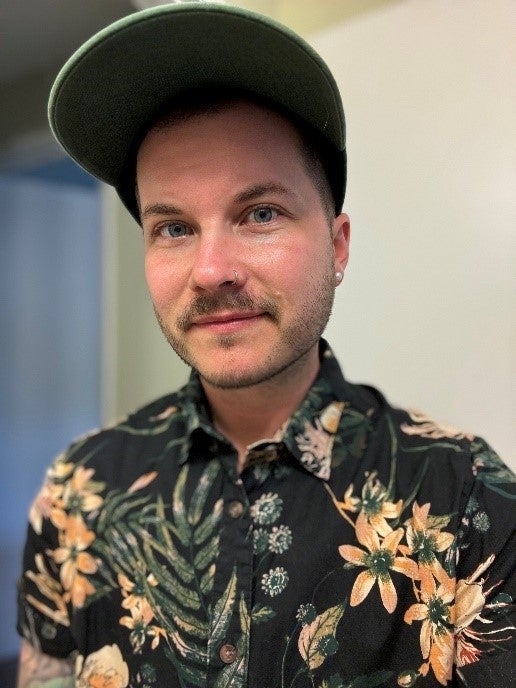 A person wearing a big green ballcap and flowered black short-sleeve shirt with an earring in their left ear sits in an office environment.