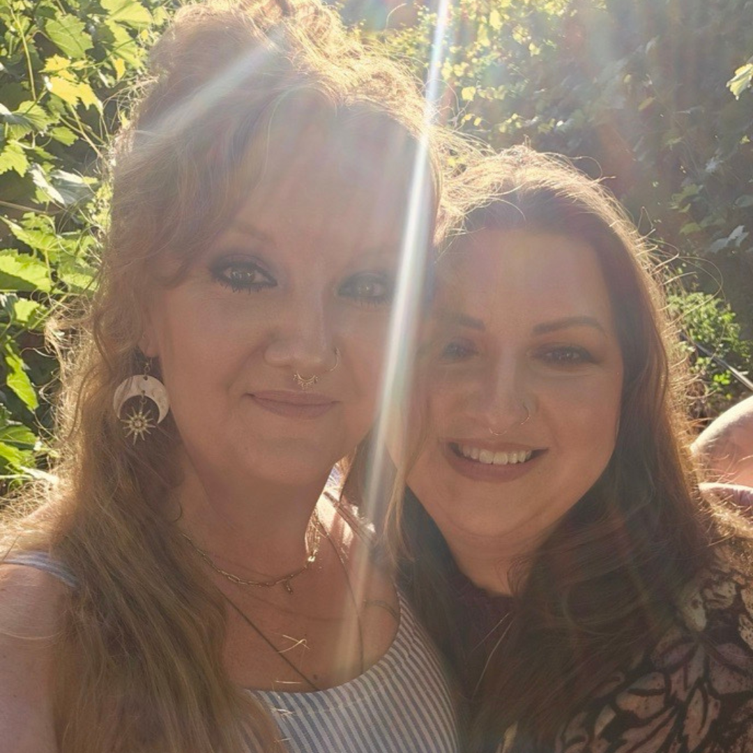 Two people standing among plants outdoors smile and face the camera while embracing each other happily.