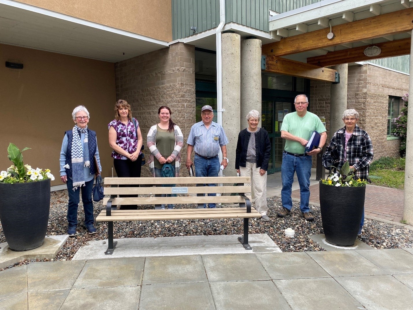 A group of seven people in various dress of different ages stand behind a wooden bench in front of a brick building. There are two flower planters either side of the group.