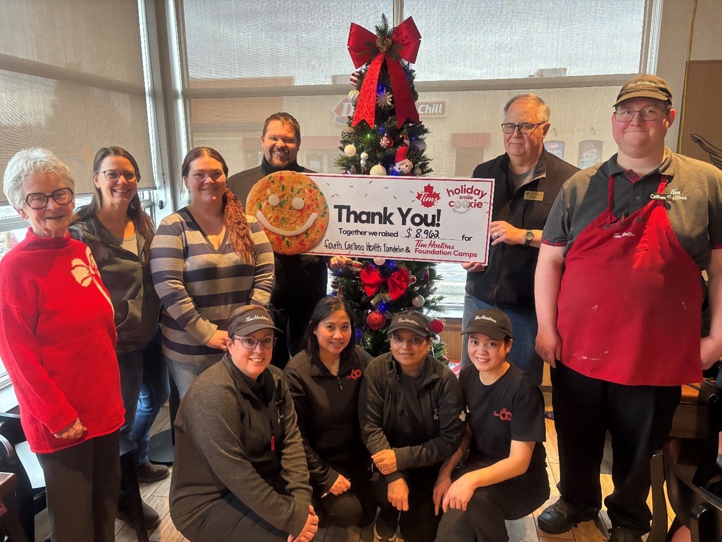 A group of 10 people wearing winter clothing and uniforms stand next to a Christmas tree. Two are holding a big cheque that says Thank You!