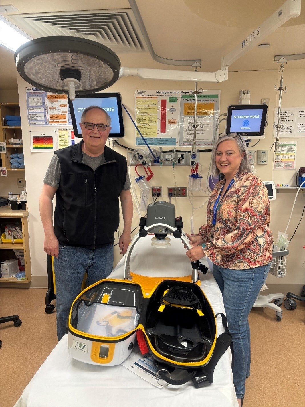 Two people with grey hair, one wearing a black vest and one a colourful blouse, stand next to a yellow and black apparatus sitting on a hospital bed. There are monitors, lights and posters behind them.