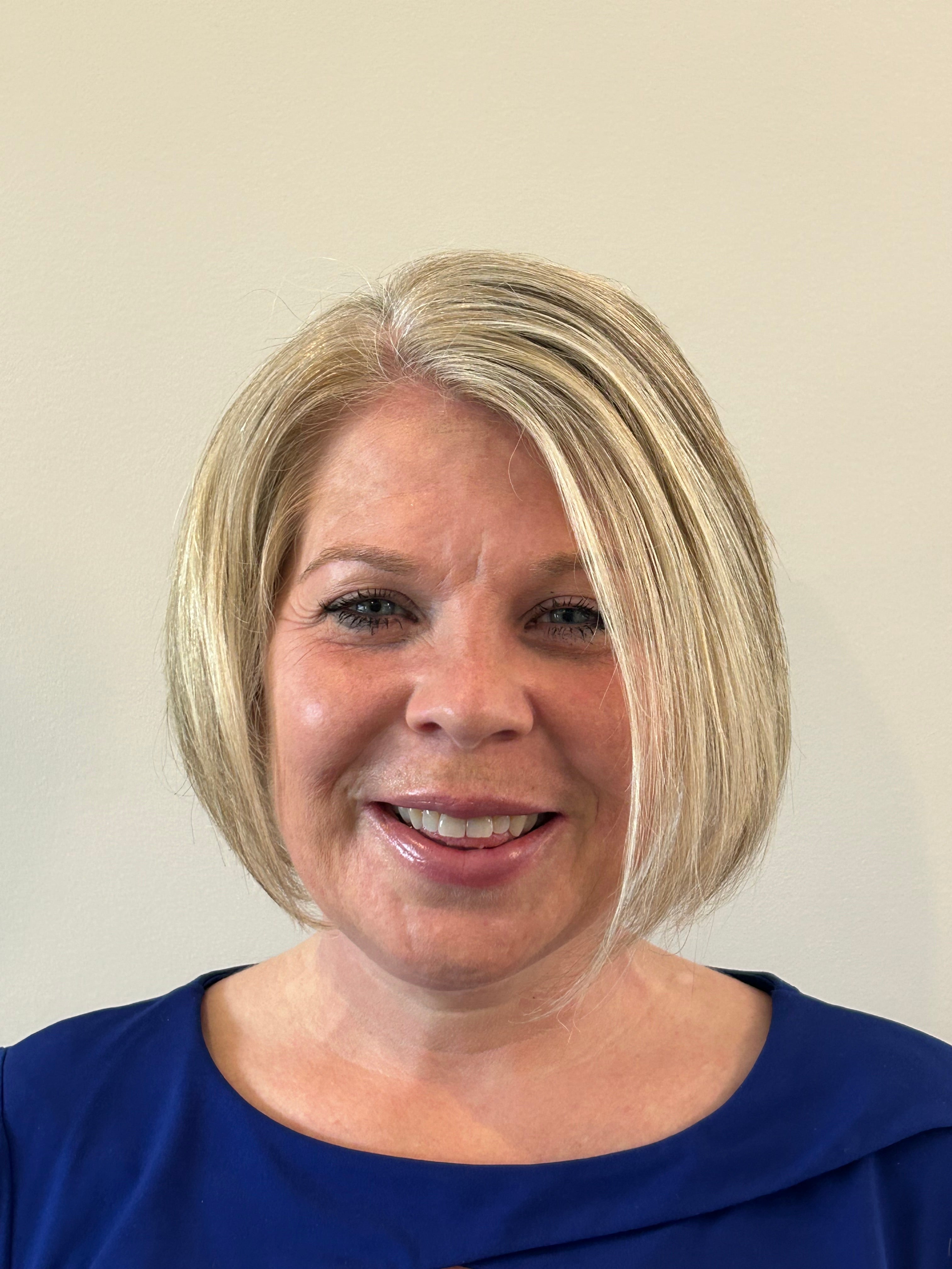 A smiling person with short blonde hair cut into a bob wearing a black shirt standing in front of a blank wall.