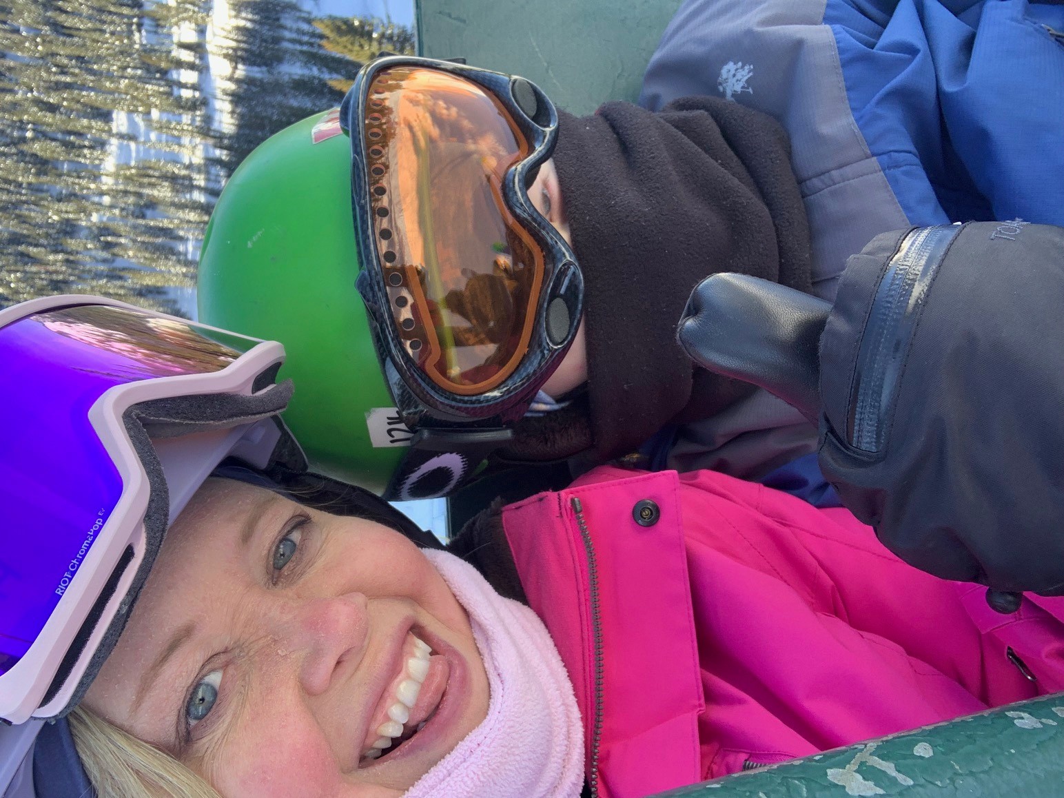 Woman and child sit on chair lift wearing goggles, helmets, and warm clothing. The woman gives a thumbs-up