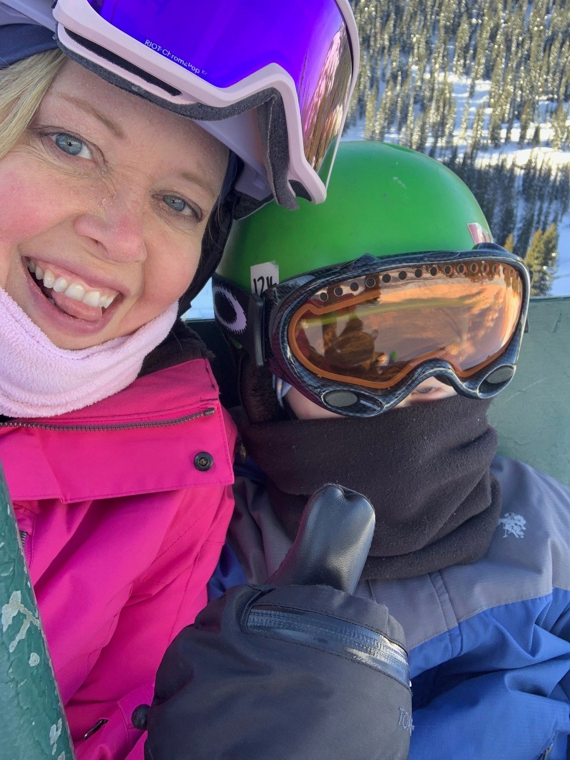 Woman and child sit on chair lift wearing goggles, helmets, and warm clothing. The woman gives a thumbs-up