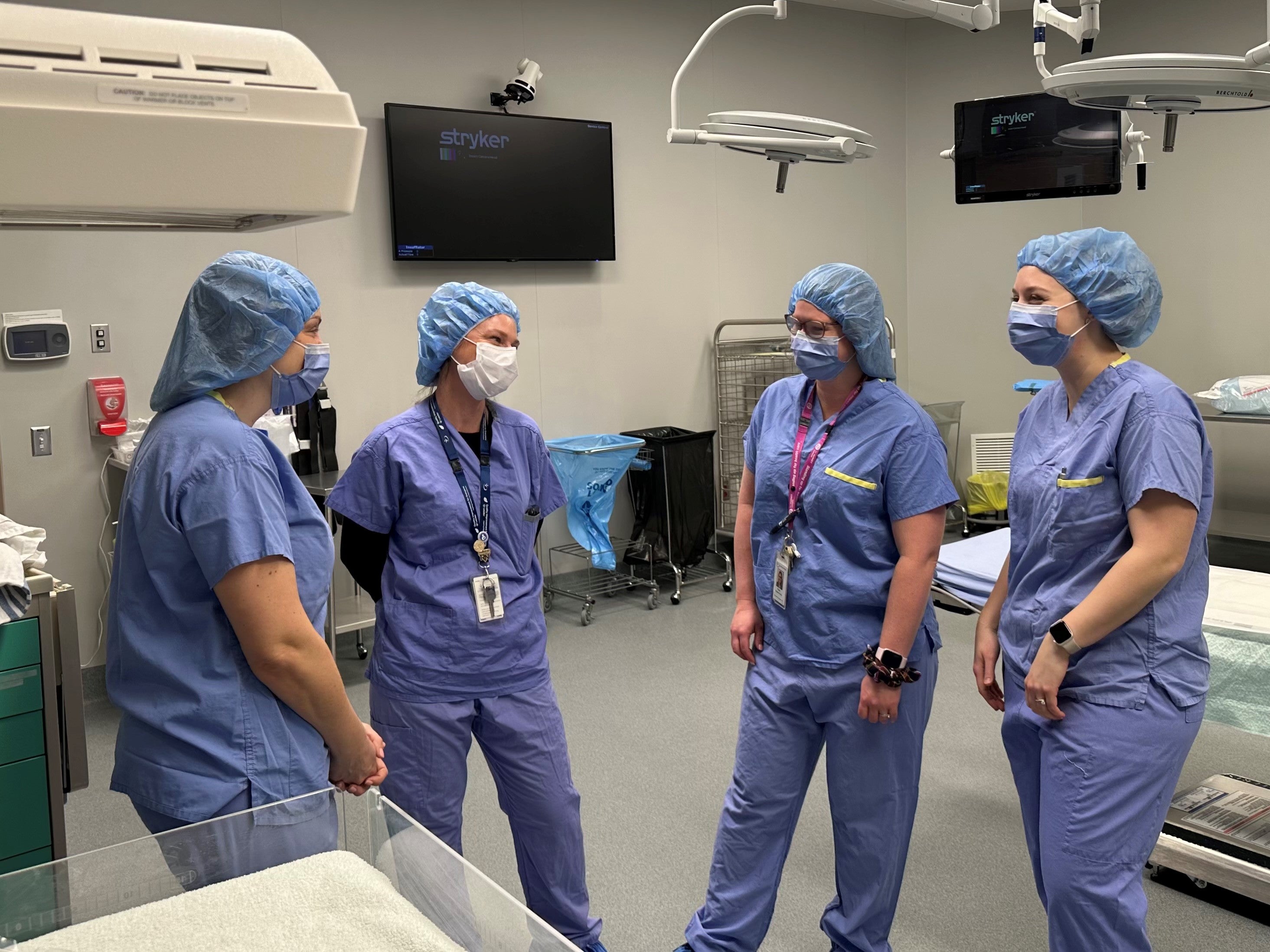 Four health-care professionals in scrubs wearing masks and head caps stand and chat in a medical room.