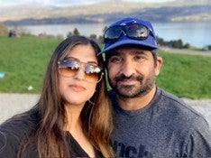 A woman with long dark hair and sunglasses smiles next to a man with a blue ball cap, a beard and grey t-shirt in front of a lake and green grass