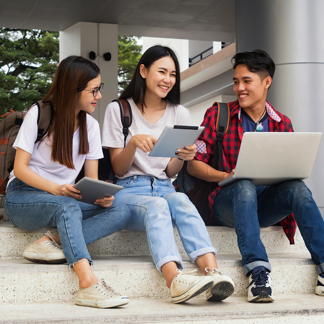 Three students sit on the steps of a school, smiling and laughing as they look at a laptop and digital notepad.