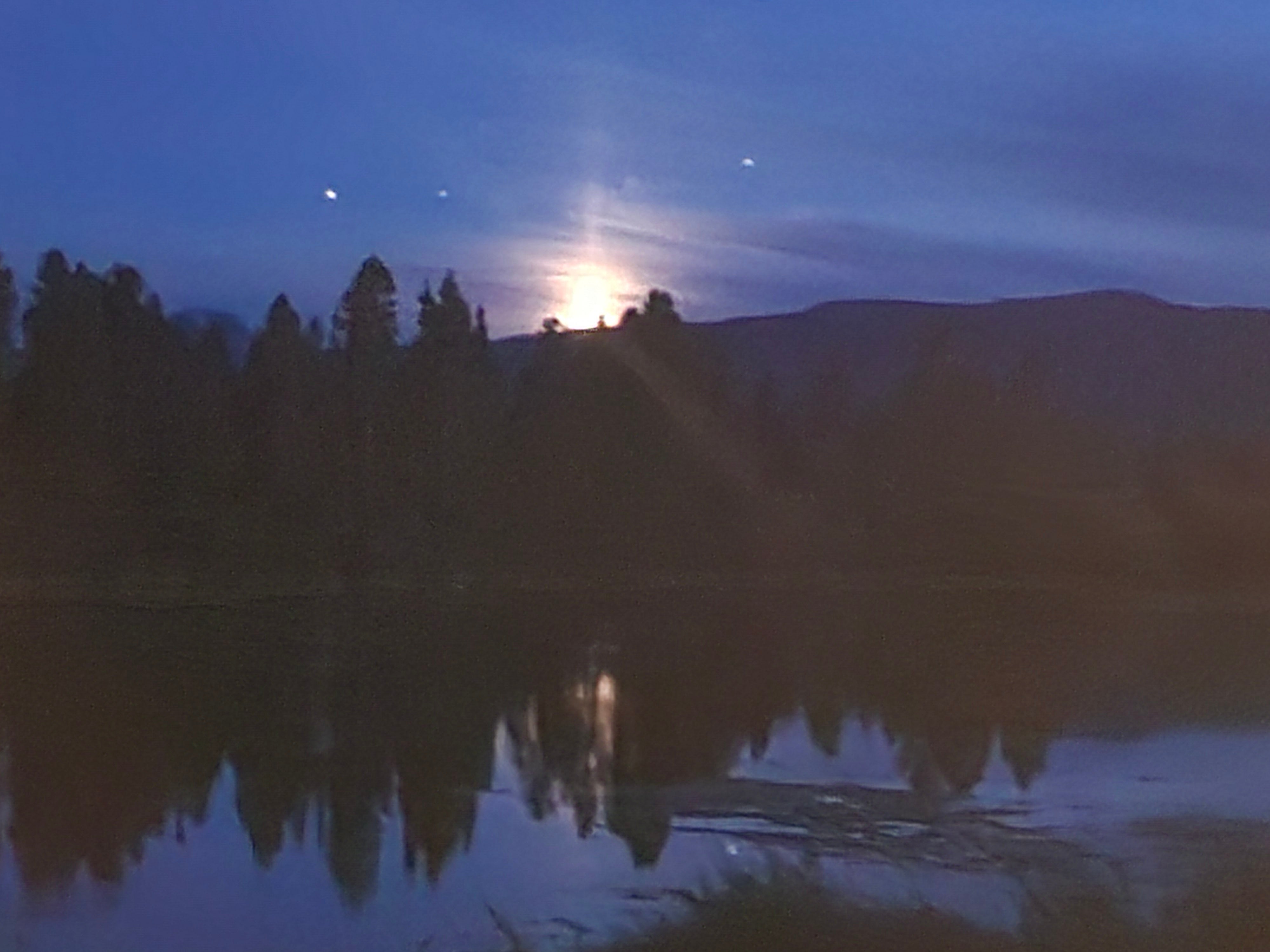 Trees reflected in a dark lake as the sun comes up over the horizon.