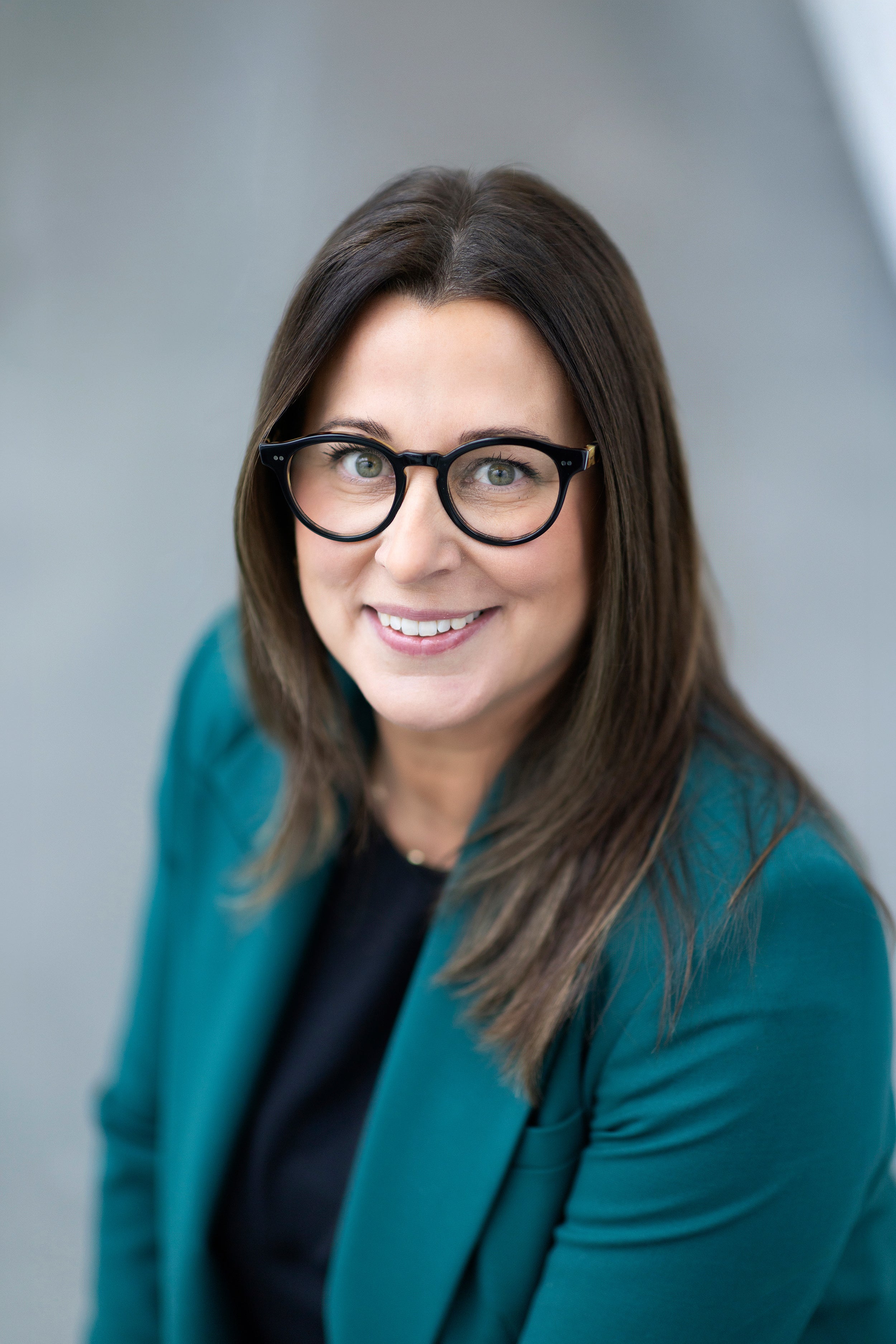 Profile shot of a smiling person with long dark hair wearing glasses, a black shirt and aqua blazer.