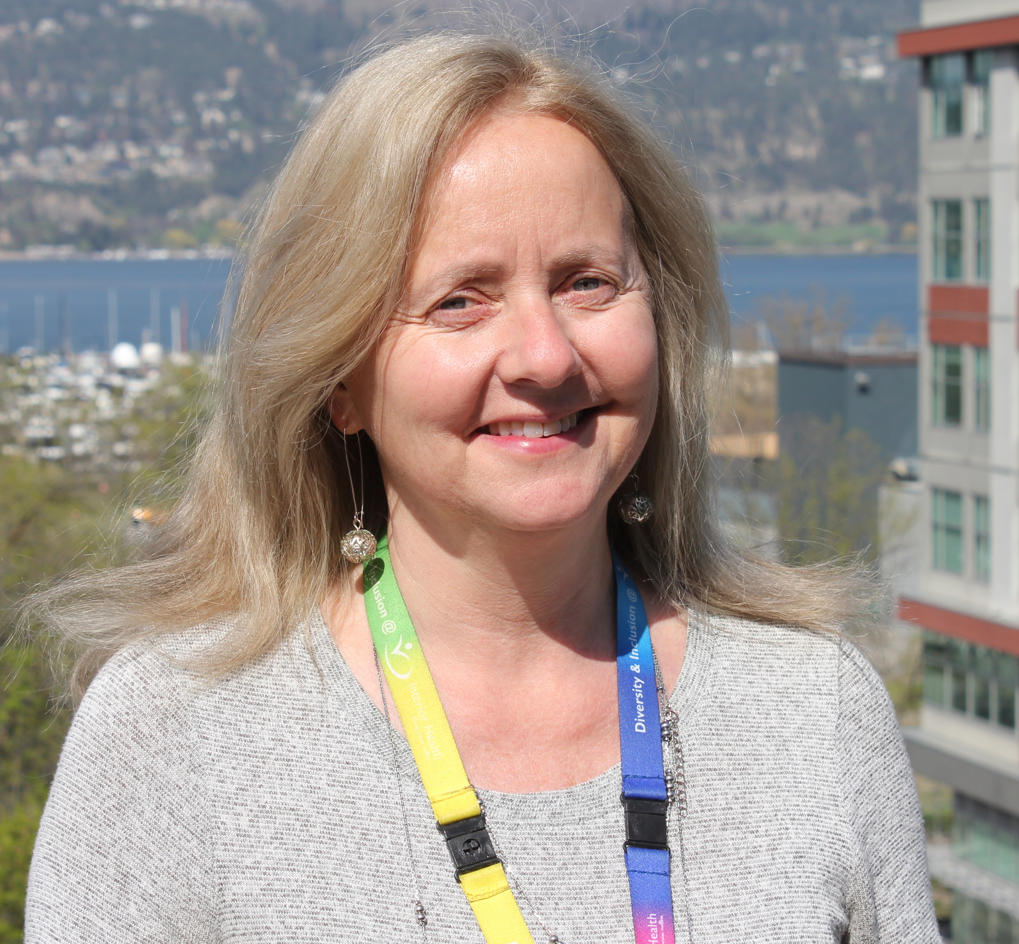 A smiling woman with blonde hair wearing a grey shirt and multi-coloured lanyard poses for a picture with a large building, water and mountains in the background.