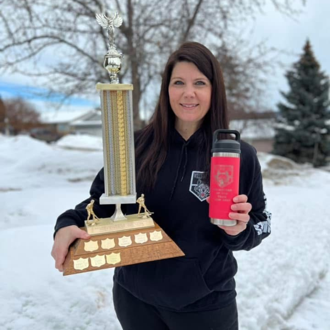 A person with long, dark wavy hair, with blue eyes wearing a black hooded sweatshirt is holding a hockey trophy while holding up their pink travel mug.
