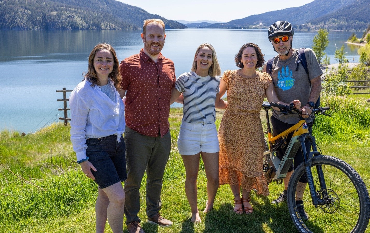 A group of five people in summer attire and one wearing a biking helmet with a bike stand arm in arm in front of a lake on a sunny day.