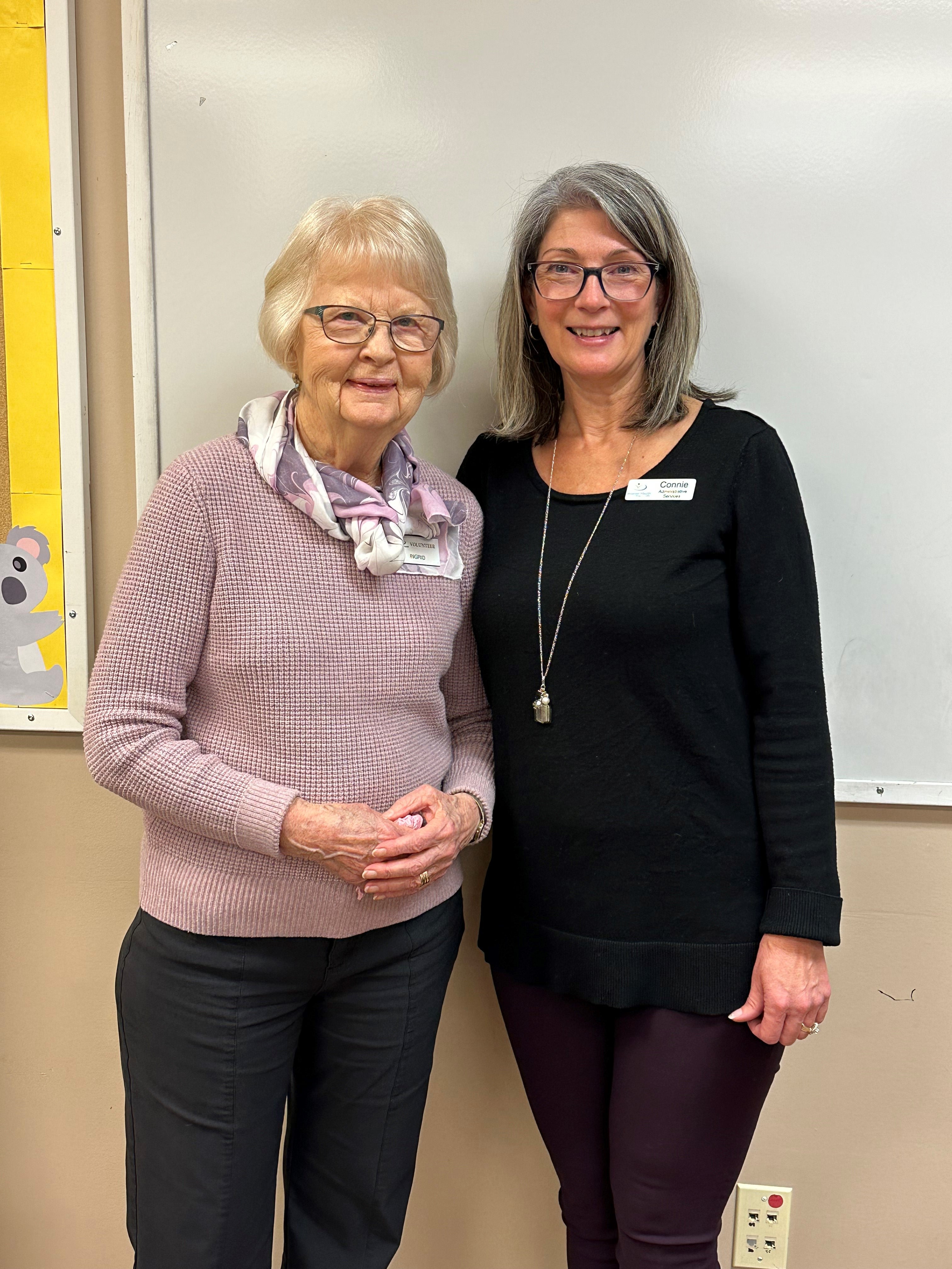 Woman with white hair, wearing glasses, lilac sweater and scarf standing beside woman with grey hair, wearing glasses, black top and long necklace.