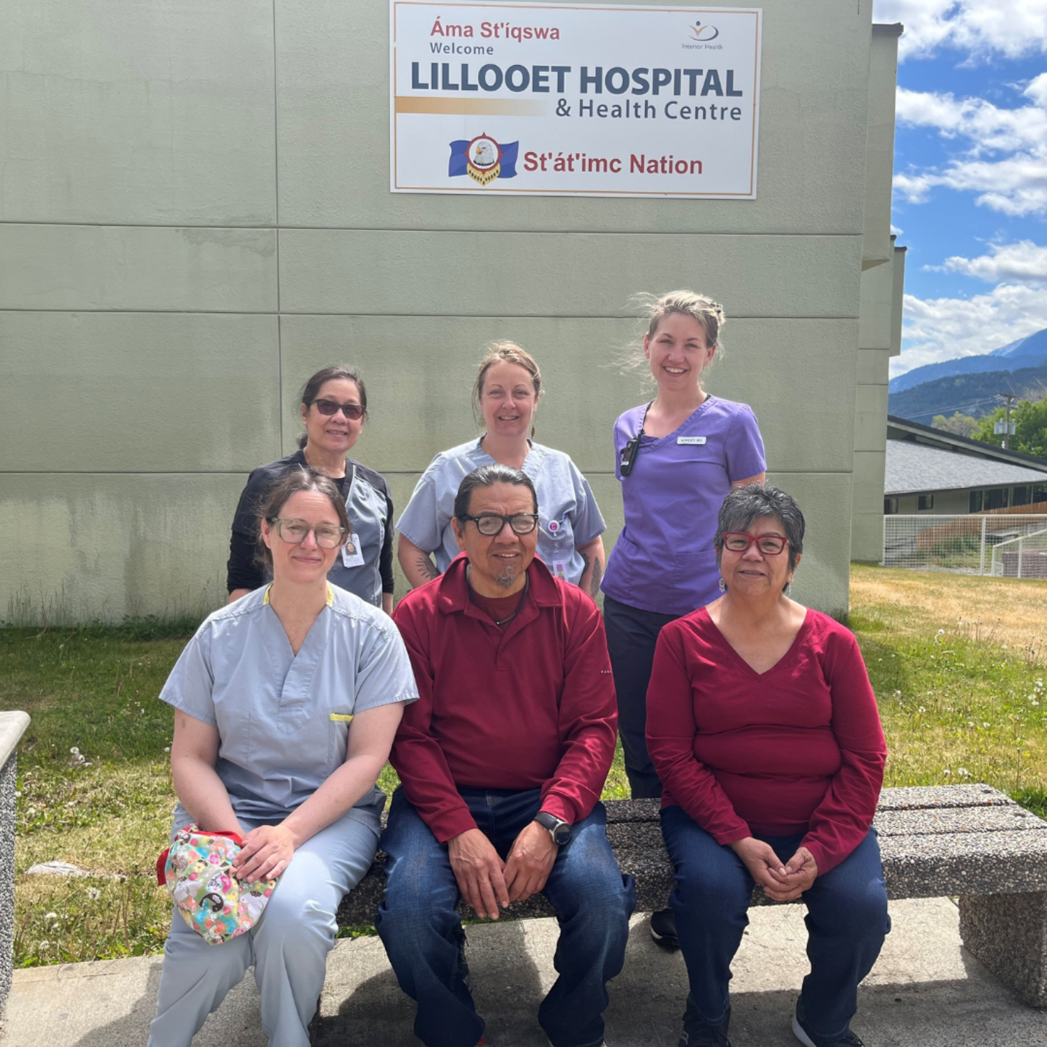 Three people seated on a bench with three people behind them standing together in front of the Lillooet Hospital & Health Centre sign on the building. 