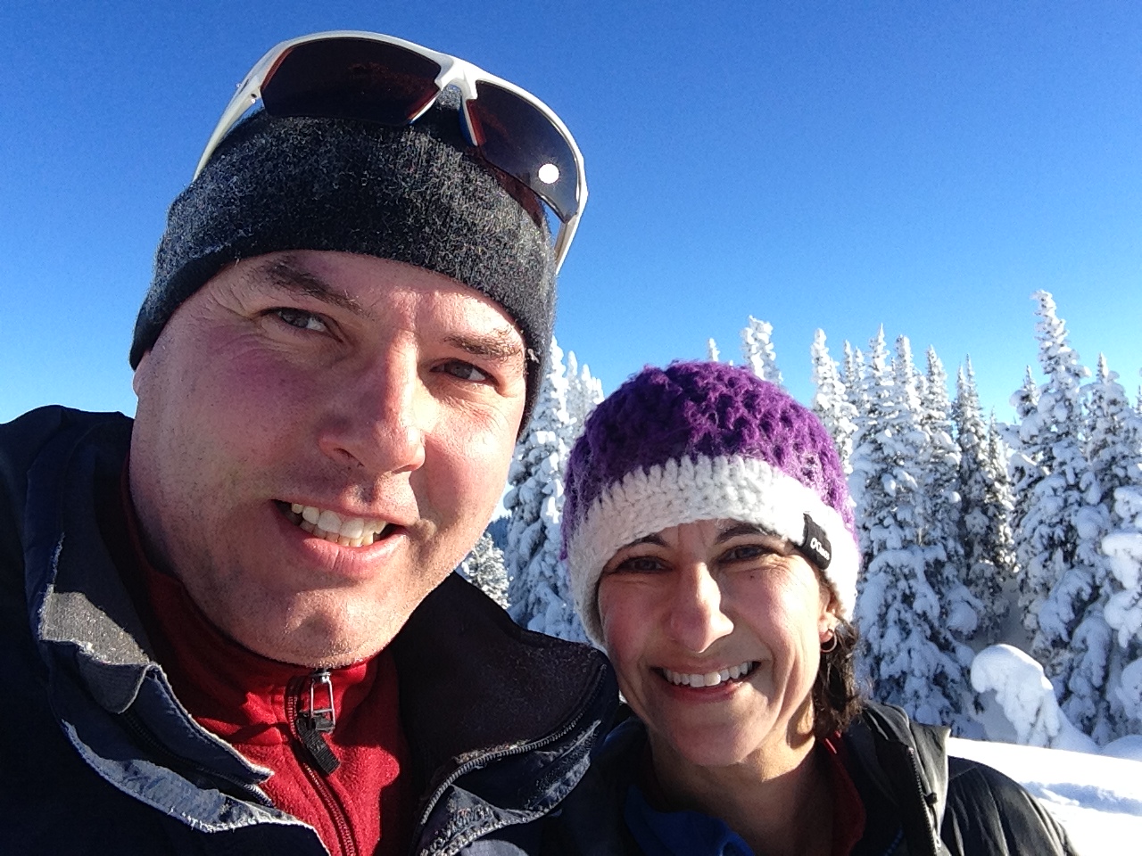 Man and woman wearing winter hats and sunglasses, hiking in snow with trees behind them.