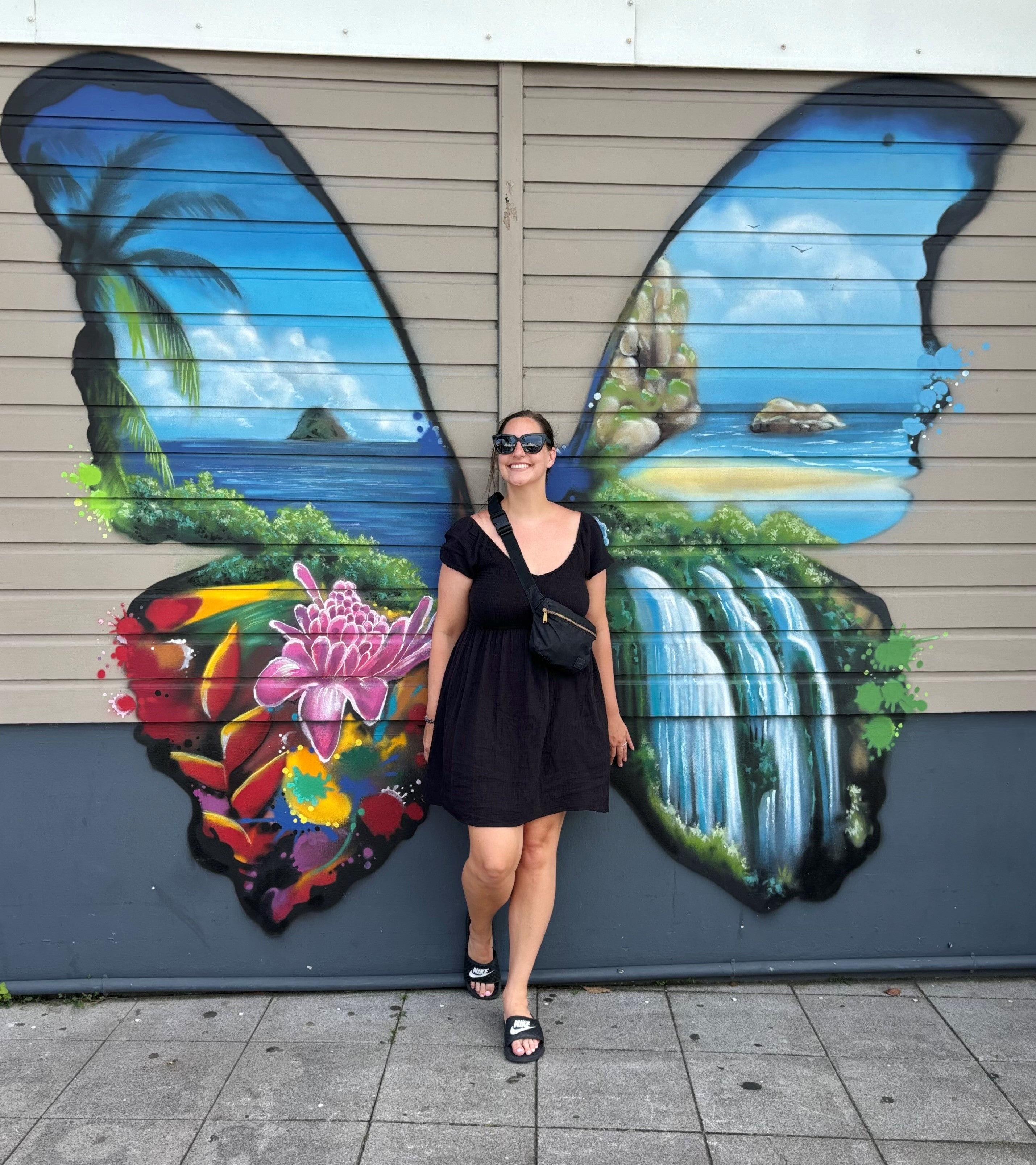Smiling woman wearing sunglasses, short-sleeved black dress and shoulder-bag, stands in centre of a wall with beige horizontal panels and solid grey lower third, with large butterfly wings painting that has ocean scenes, flowers and a waterfall within the wings.