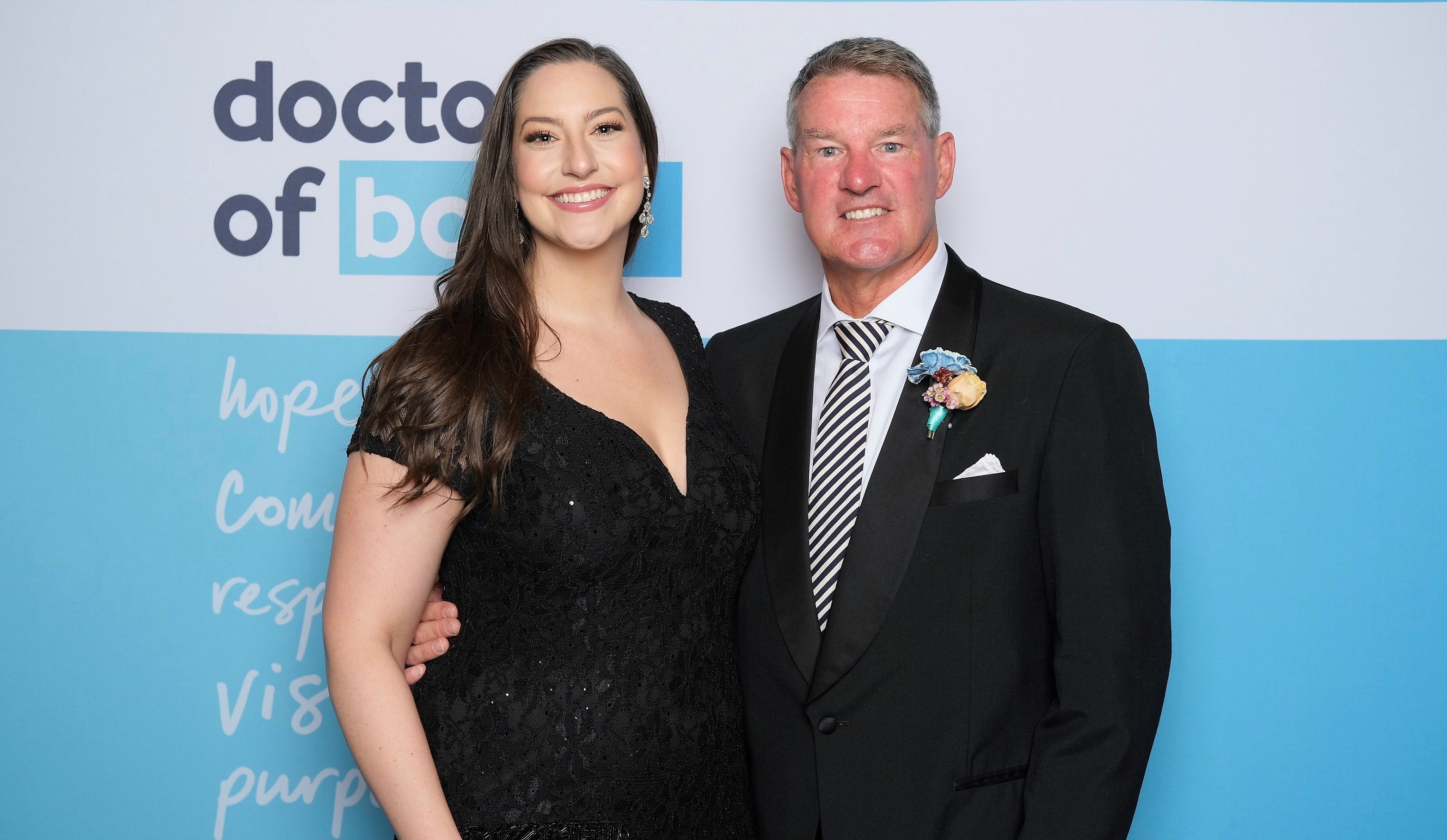Smiling woman with long brown hair wearing black lace v-neck dress stands next to grey-haired man in black suit with white shirt, black and white diagonal striped tie, with light blue and white wall behind them.