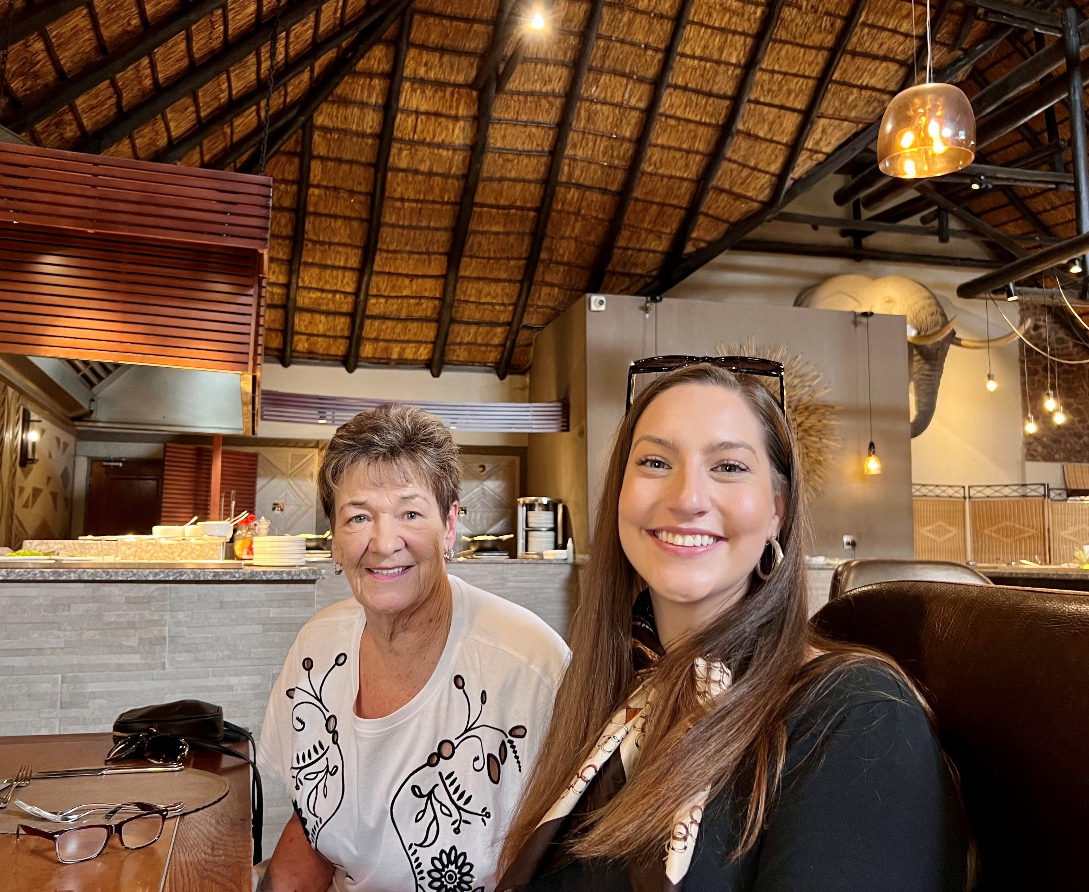 Two smiling women sitting together with older, short-haired woman on left wearing white sweater with black floral design, and younger, long brown-haired woman on right wearing sunglasses on top of head and black top, with high wood ceiling and hanging lamps in background.
