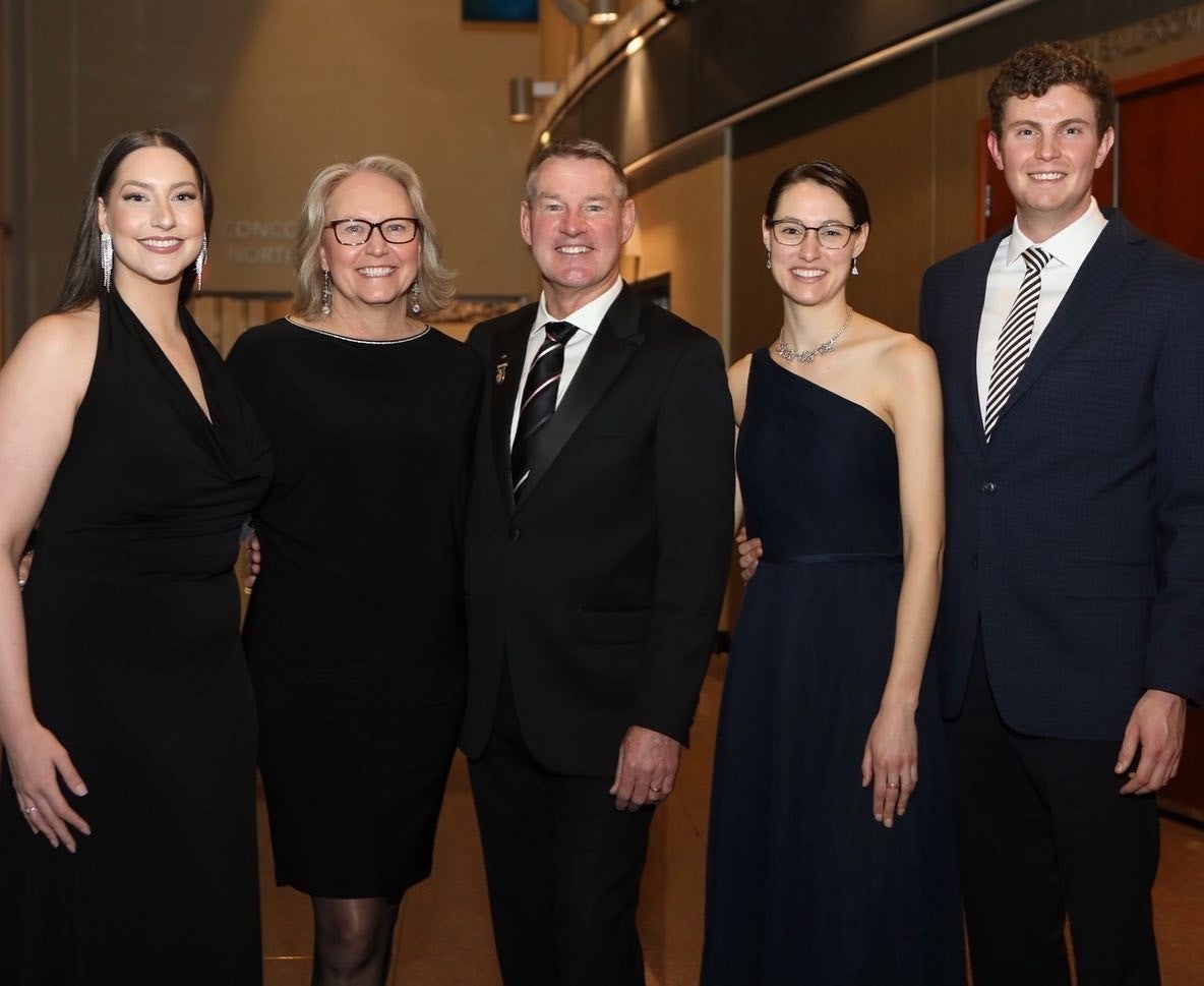 Five smiling people, three women and two men, stand shoulder-to-shoulder wearing formal black with beige and brown wood walls behind them.