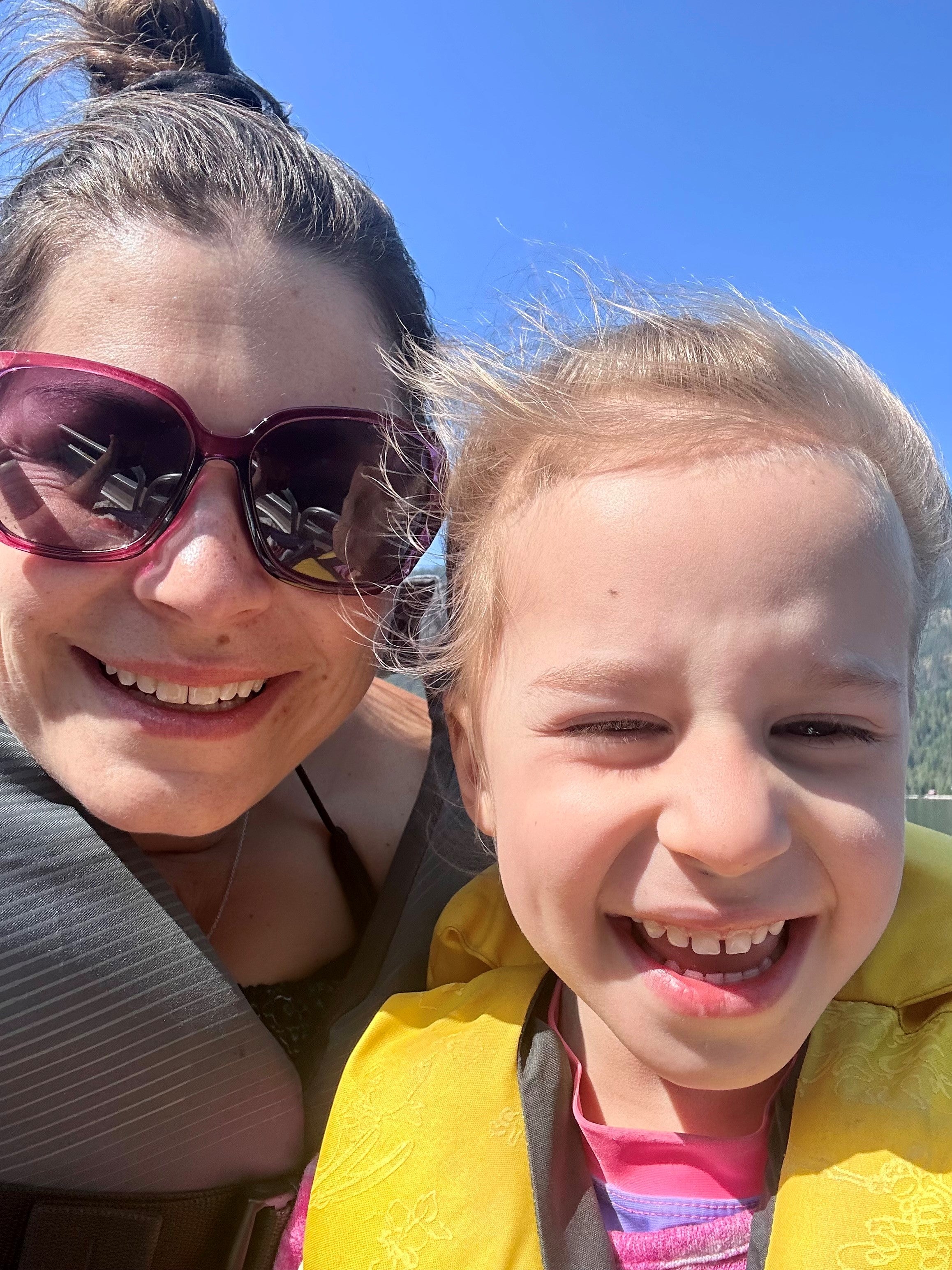 Smiling woman with brown hair and large sunglasses with red frames, with smiling, squinting blonde girl wearing yellow life jacket and pink top