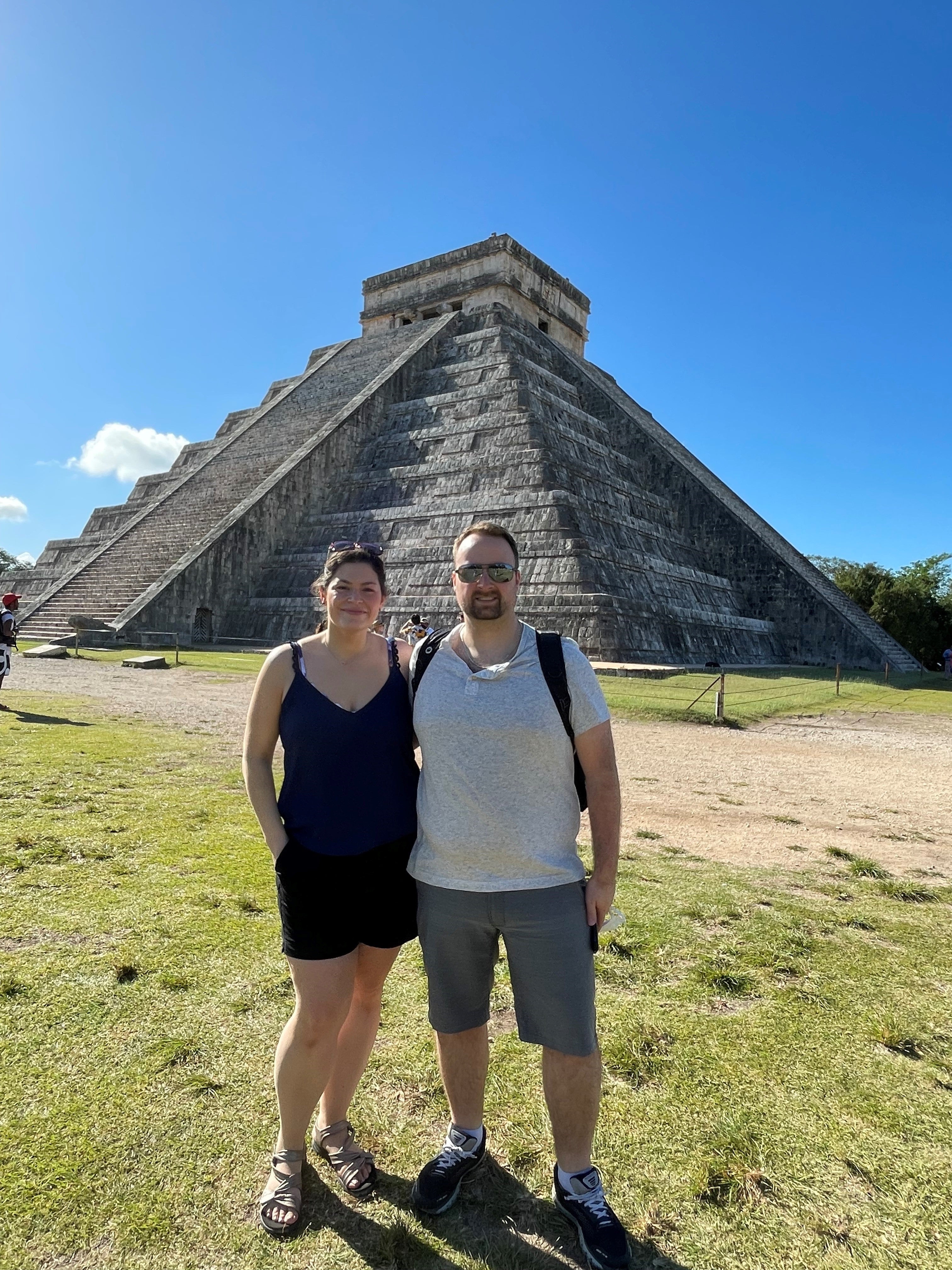 Smiling woman with brown hair wearing sleeveless black top and black shorts standing next to man with beard and moustache wearing sunglasses, white short-sleeved shirt, shorts and running shoes, with green grass, blue sky and ancient pyramid structure with stairs in the background