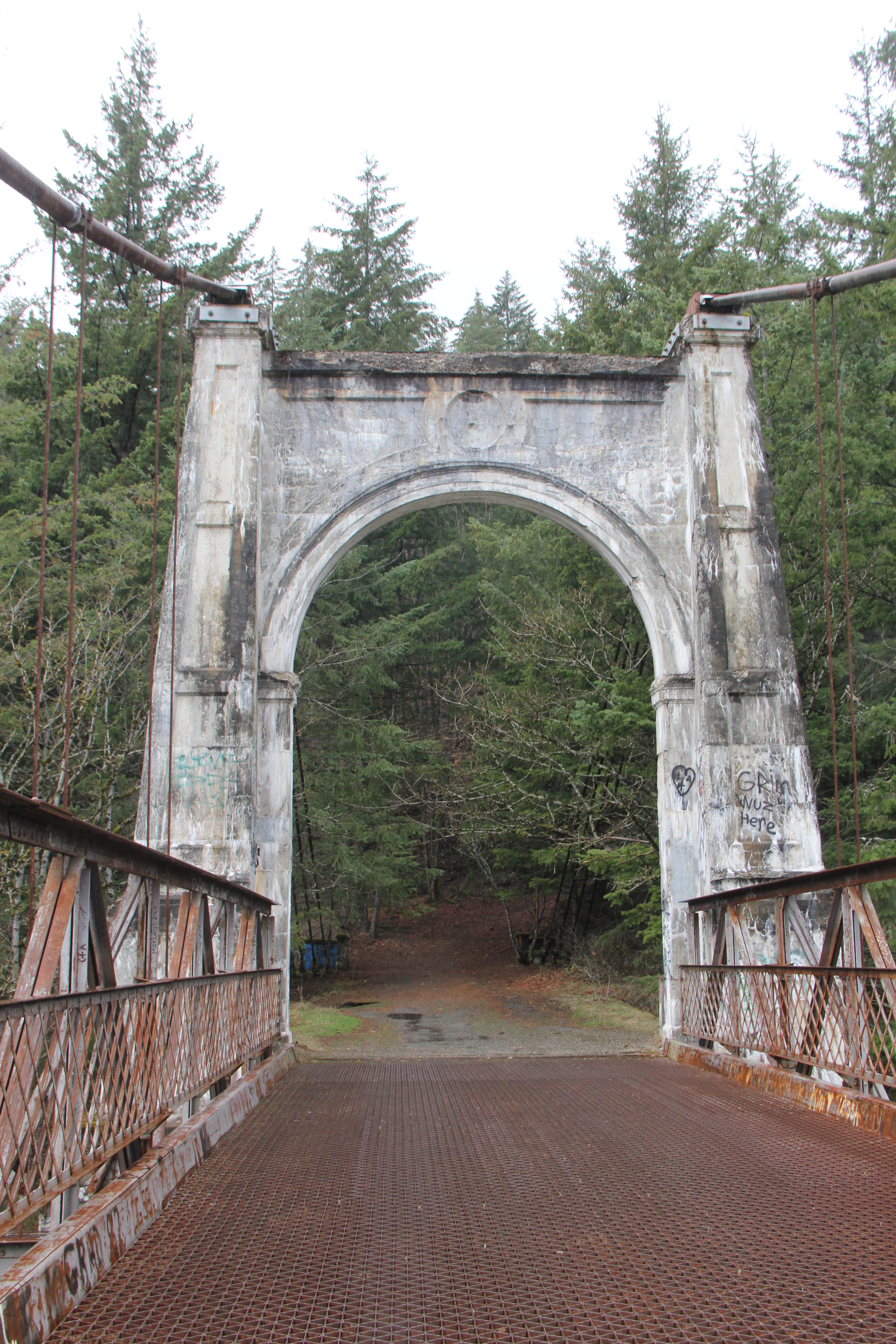 A bridge with a large supporting white arch leads into a forested area.