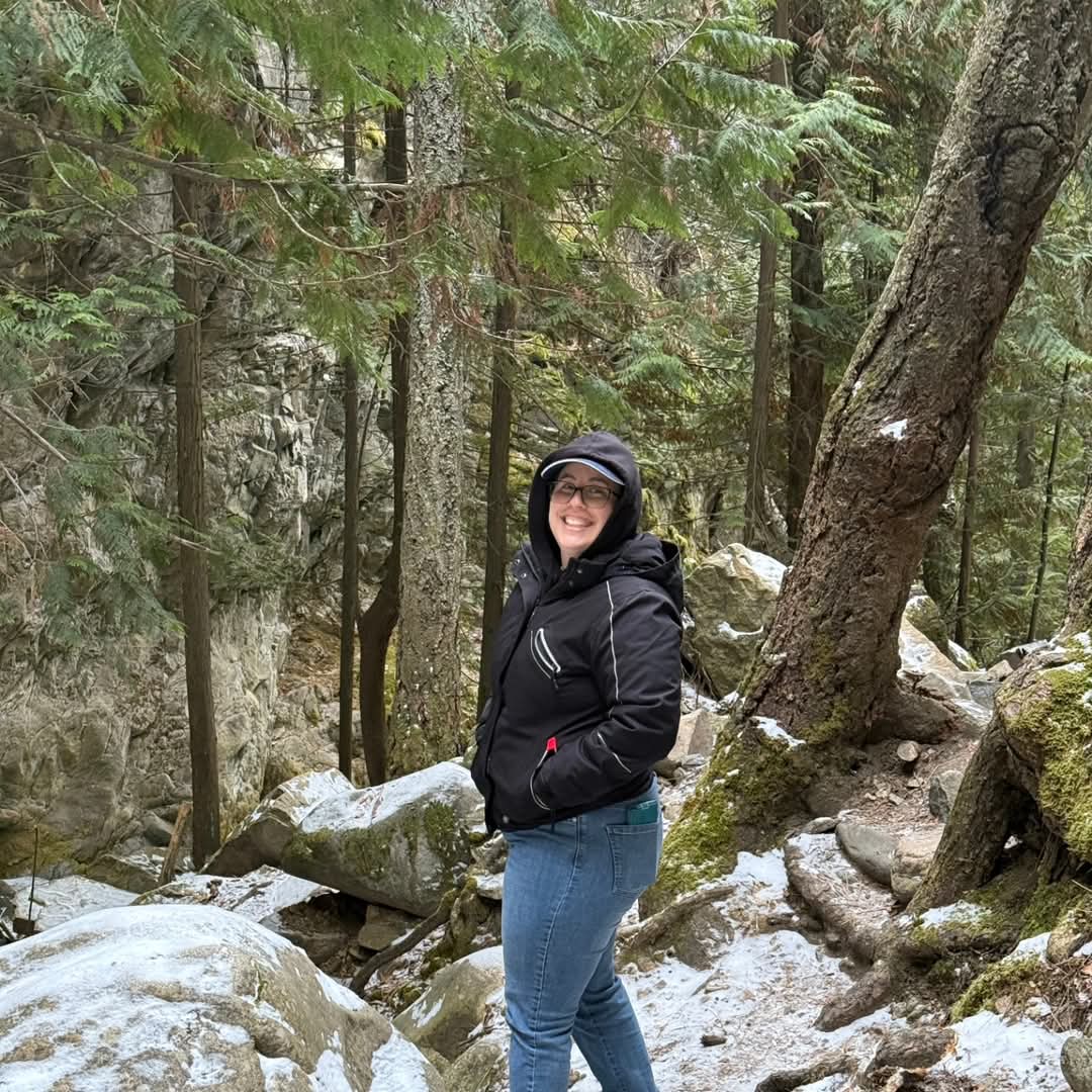 A woman in a black, hooded jacket wearing a ball cap is in snow dusted woods and turns around to smile at the camera.